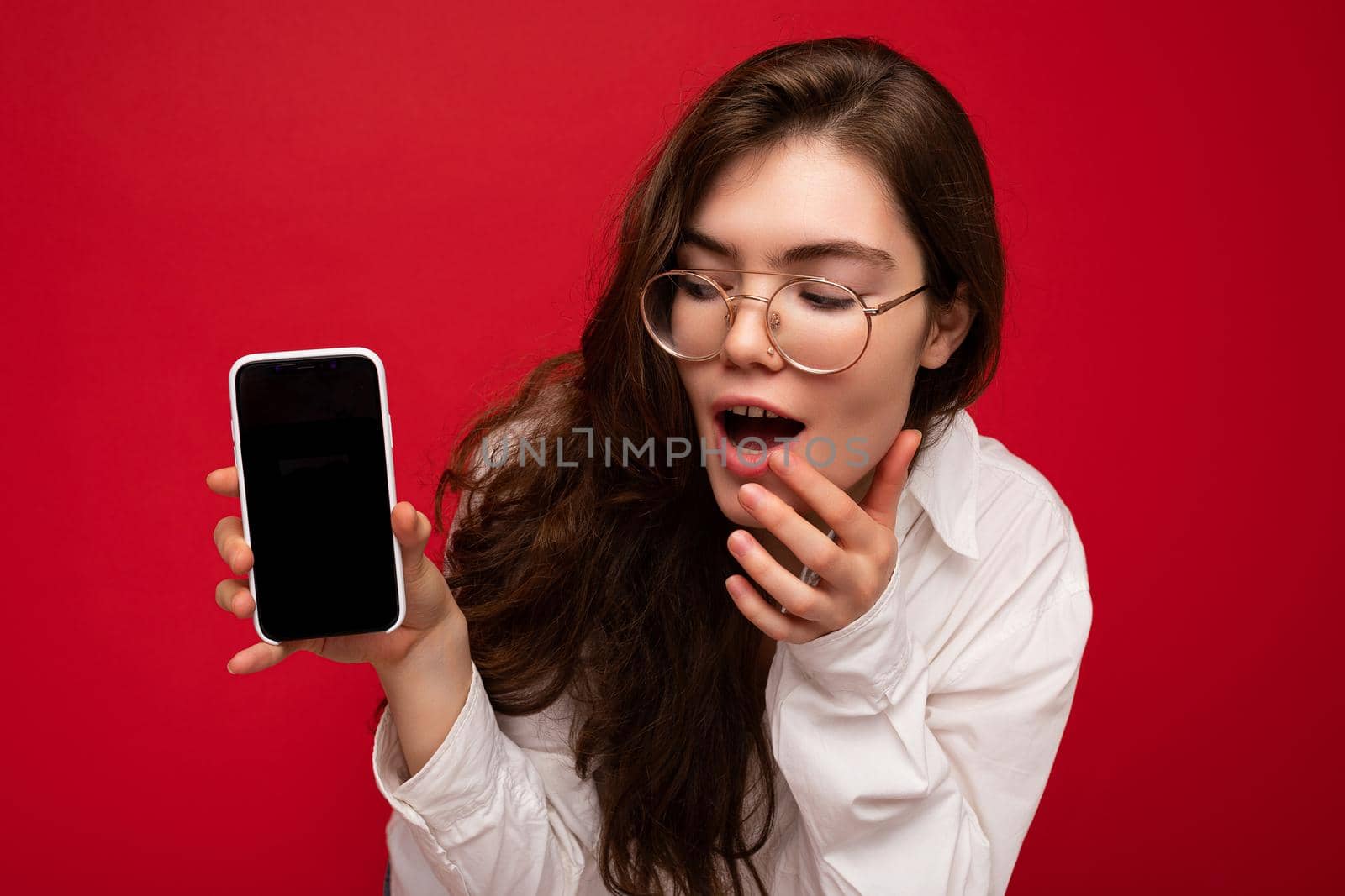 photo of sexy amazed beautiful young brunette woman wearing white shirt and optical glasses isolated over red background holding in hand and showing mobile phone with empty screen for cutout looking at gadjet screen.