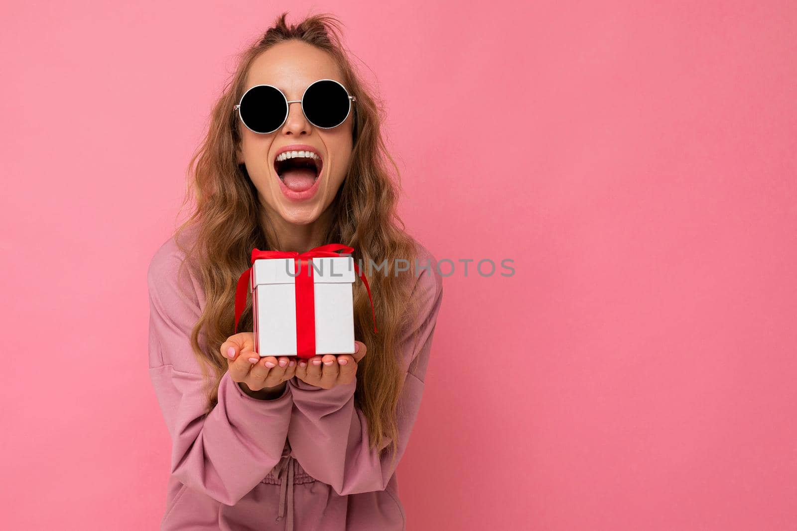 Photo shot of pretty positive surprised young blond woman isolated over colourful background wall wearing trendy outfit look holding gift box and looking at camera. Copy space, mockup