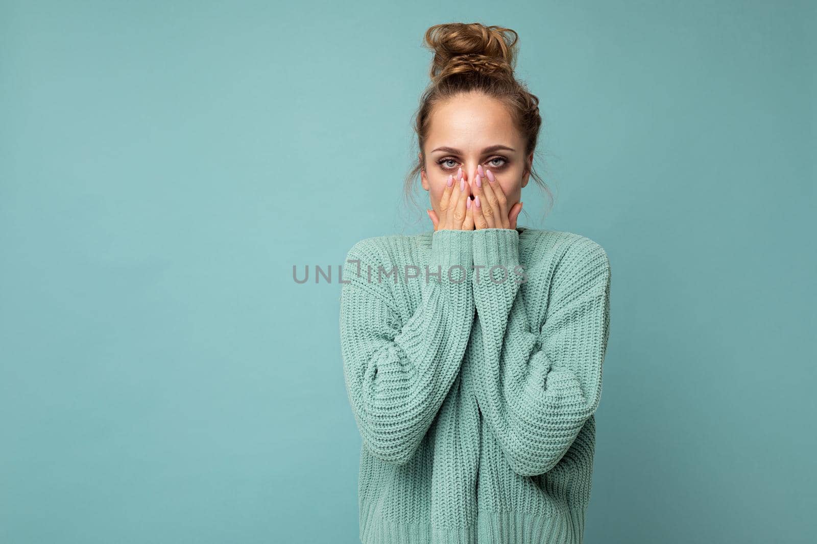 Portrait of young shocked nice winsome blond woman with sincere emotions wearing trendy blue sweater isolated over blue background with free space and covering mouth.