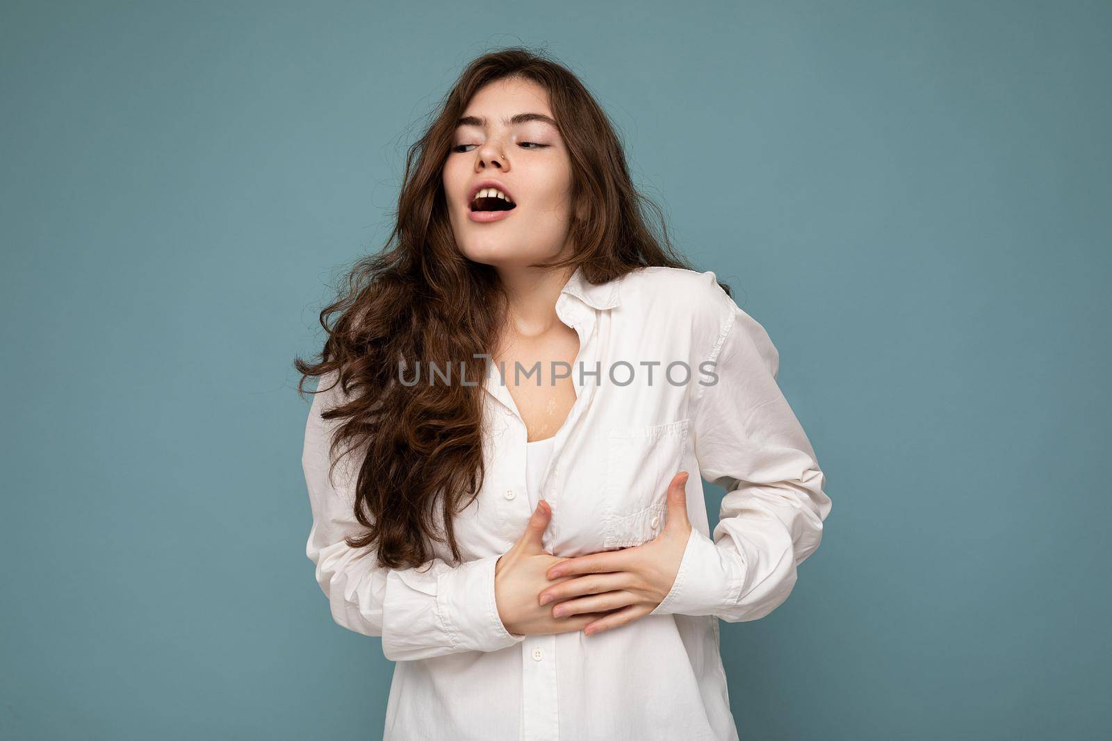 Portrait of young emotional shocked beautiful wave-haired brunette woman with sincere emotions wearing casual white shirt isolated on blue background with empty space and touches chest with hands.