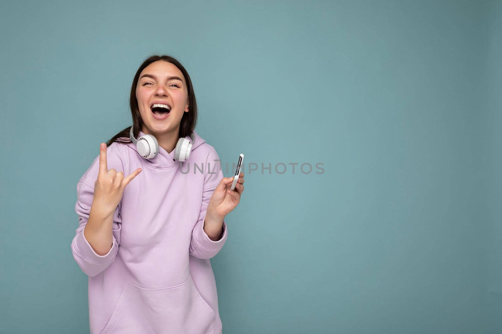 Photo of beautiful positive smiling young brunet woman wearing purple hoodie isolated on blue background holding and using smartphone wearing white wireless earphones having fun and showing rock and roll gesture by TRMK
