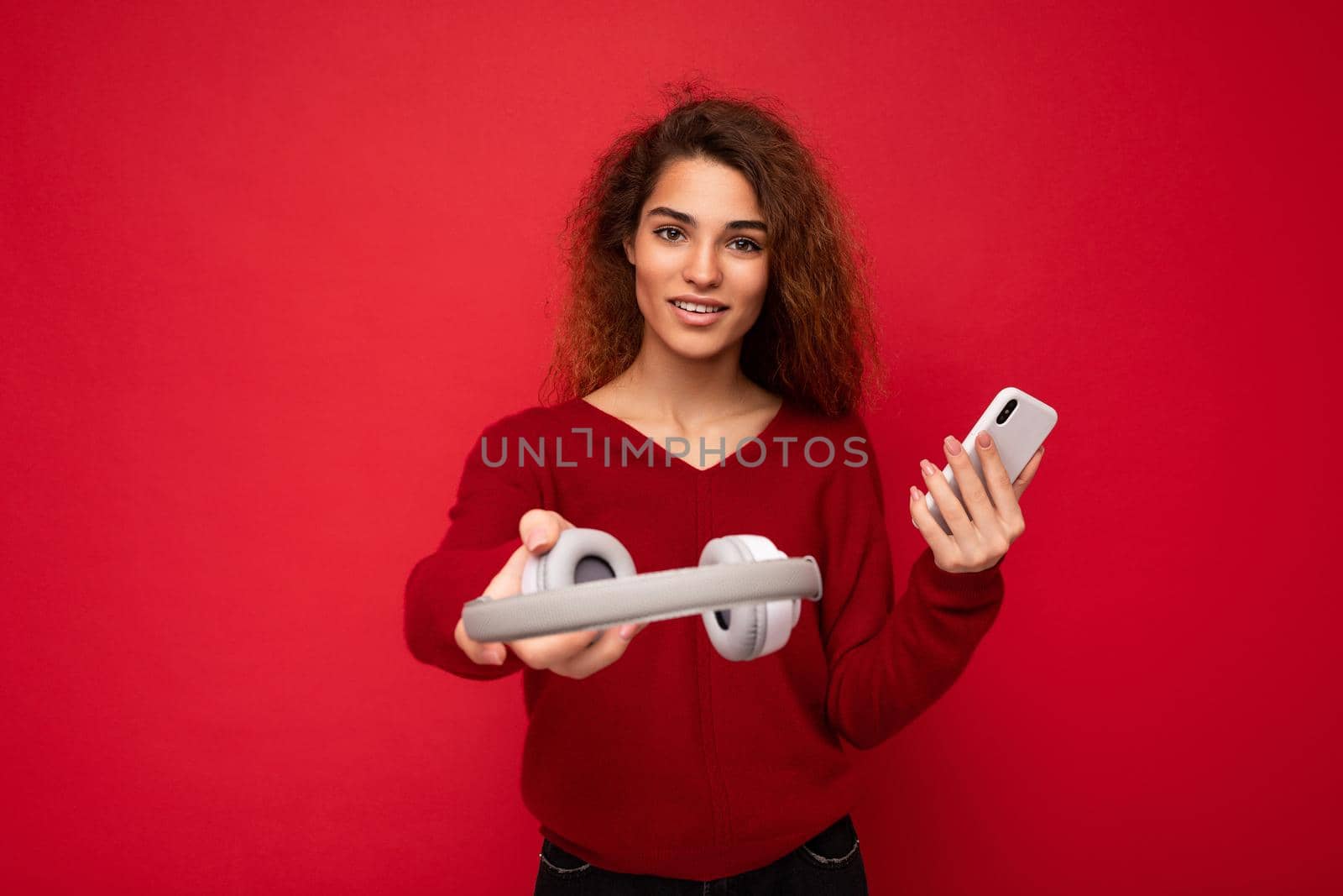 Charming funny young brunette curly woman wearing dark red sweater isolated on red backdrop holding and suggesting white wireless headphones and mobile phone looking at camera.