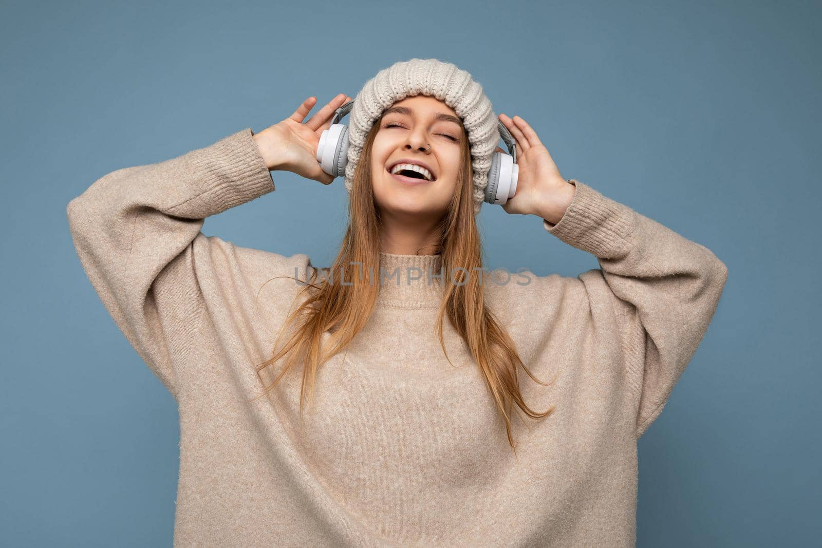 Photo of beautiful happy smiling young blonde woman wearing beige winter sweater and hat isolated over blue background wearing white wireless bluetooth headphones listening to cool music enjoying with close eyes.
