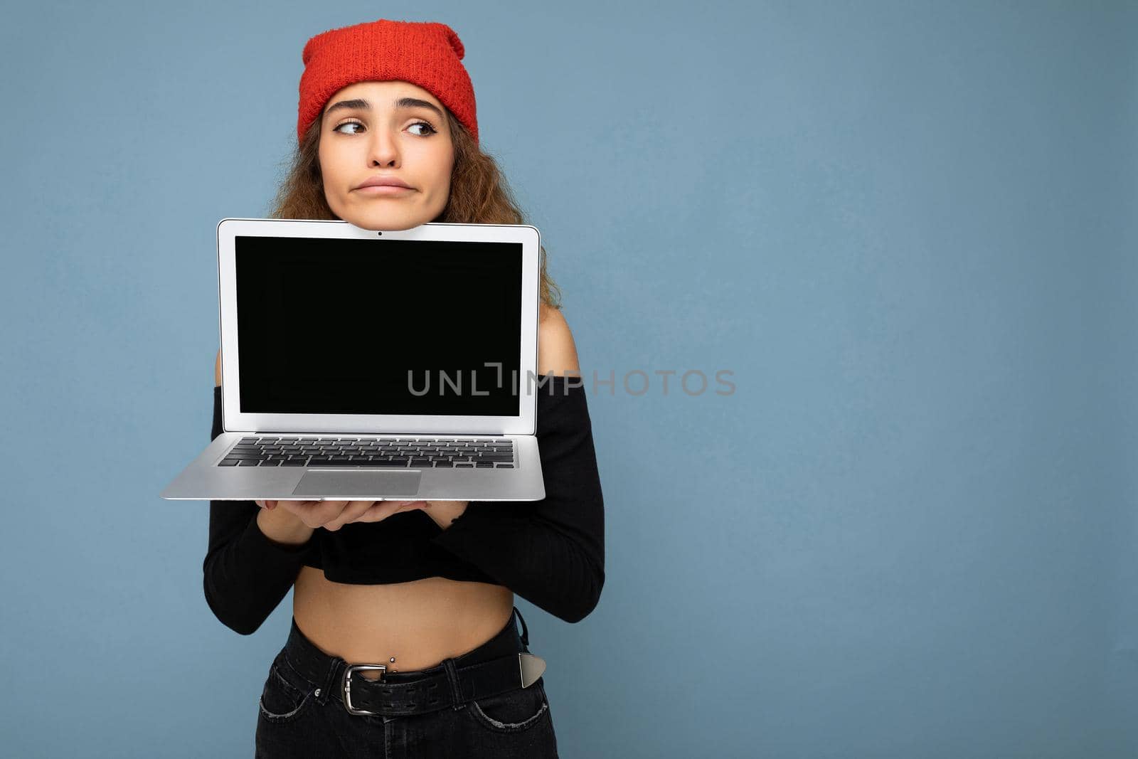 Beautiful funny amusing young brunet curly girl wearing black crop top and red and orange do-rag isolated over light blue wall background holding computer laptop with empty screen monitor looking to the side and having doubts. Mock up, copy space, empty space