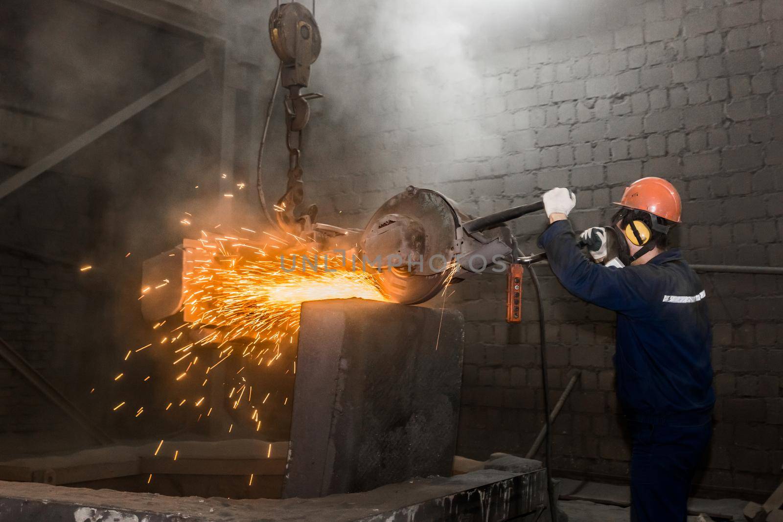 A male worker in a protective helmet, respirator, overalls manages heavy grinding equipment for cast iron concrete tubing with flying sparks in the workshop of an industrial plant.