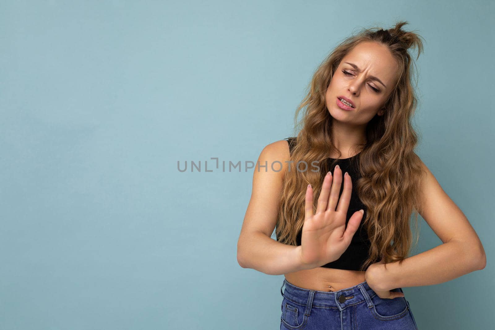 Young dissatisfied sad attractive winsome blonde curly woman with sincere emotions wearing trendy black top isolated over blue background with free space and showing palm. Negative concept.