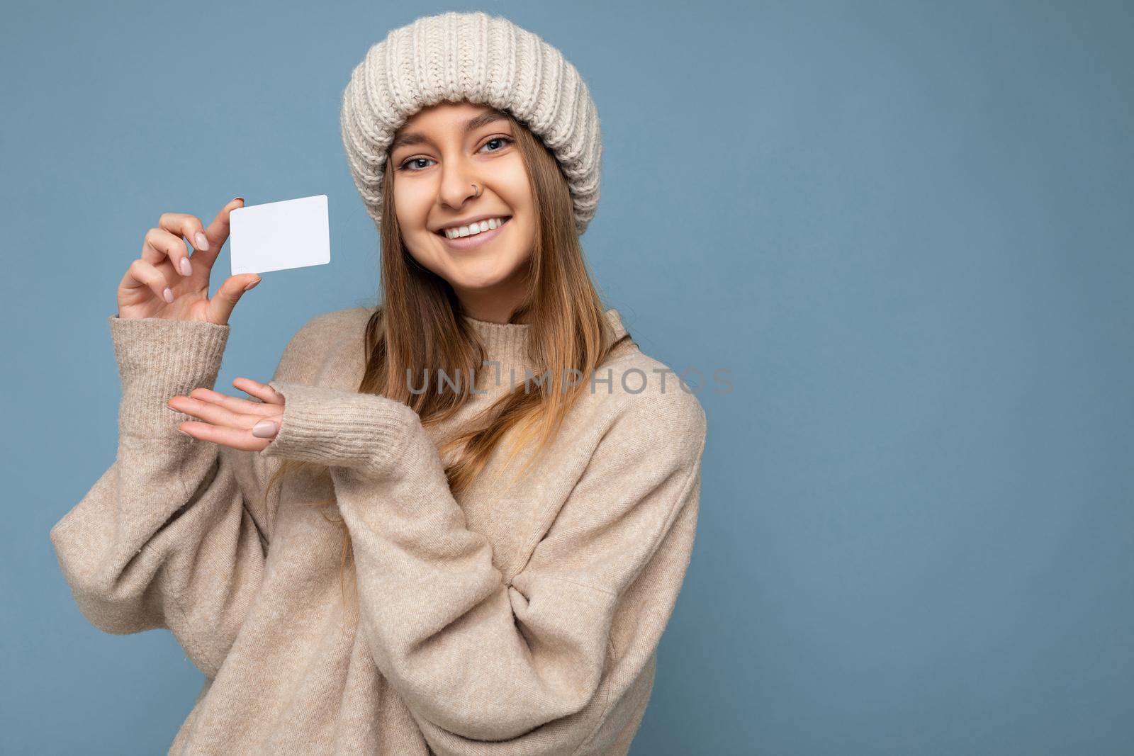 Photo shot of attractive positive smiling young dark blonde woman wearing beige sweater and knitted beige hat isolated over blue background holding and showing credit card looking at camera. copy space