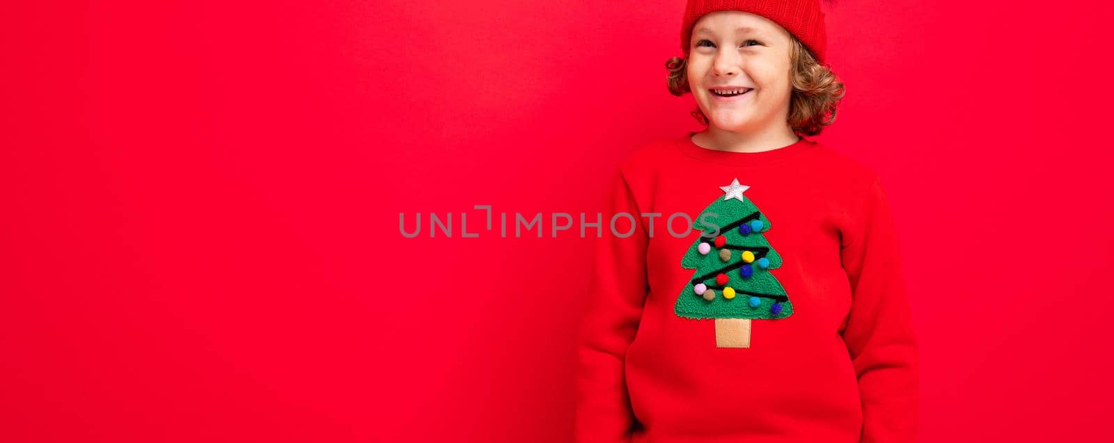 cute blond boy in warm hat and christmas sweater on red background with smile on his face.