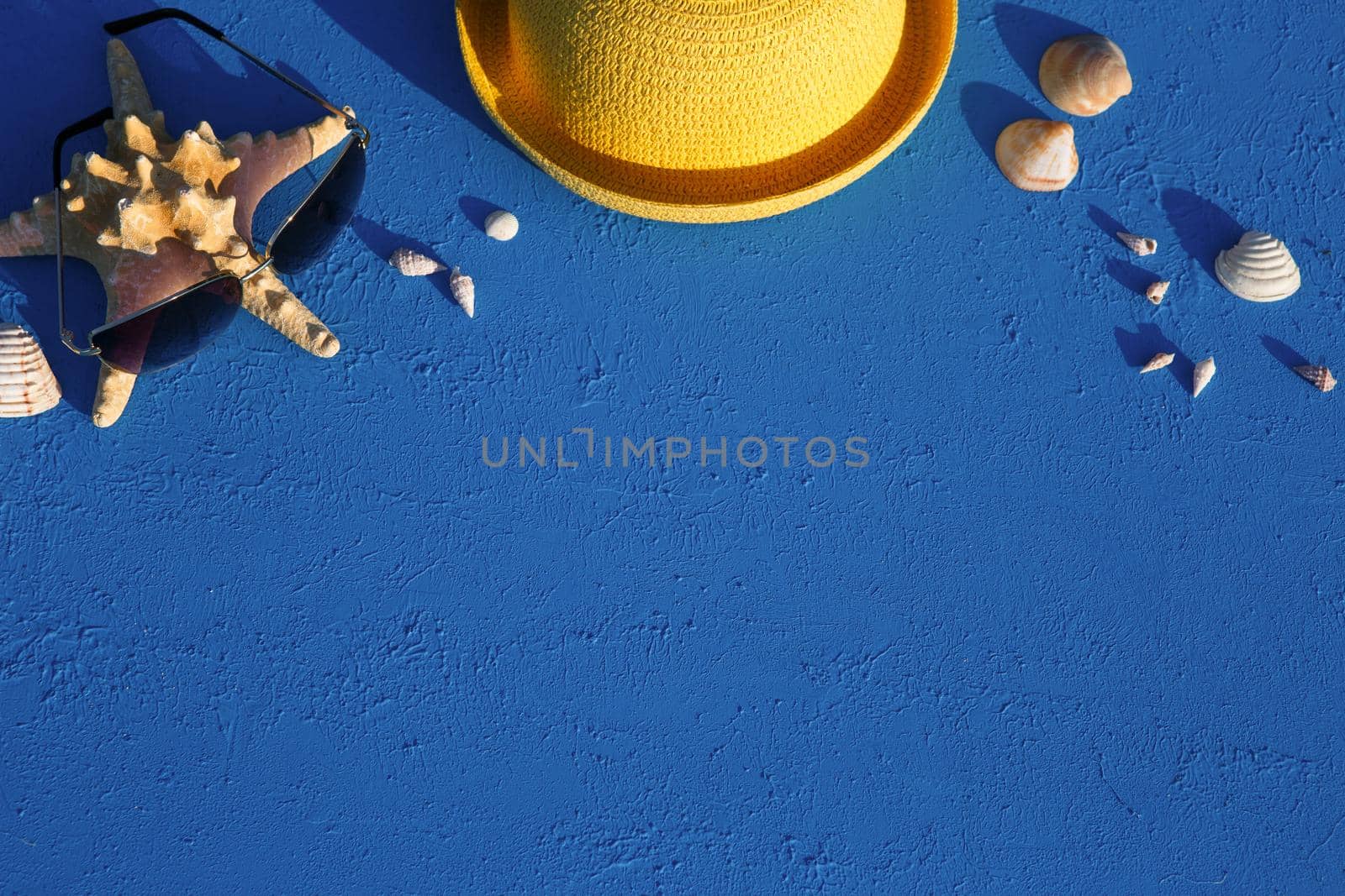Frame with beach accessories on a nautical theme: yellow straw hat, sunglasses, starfish and shells on a blue background. Vacation concept, sea trip, UV protection, swimming. Copy space. Flatlay by Simol