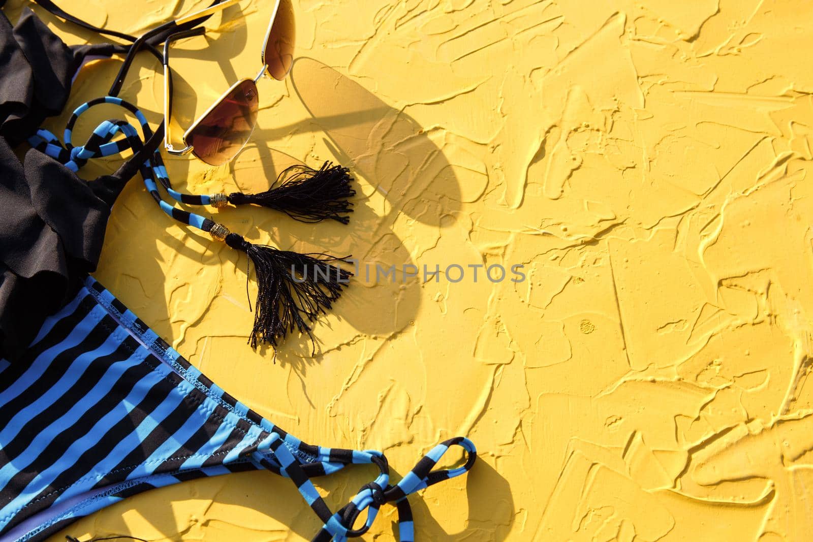 Blue with black stripes female swimsuit and sunglasses on a yellow background. The concept of a beach holiday, a trip to the sea, accessories for swimming, UV protection, tanning. Copyspace. Flatlay