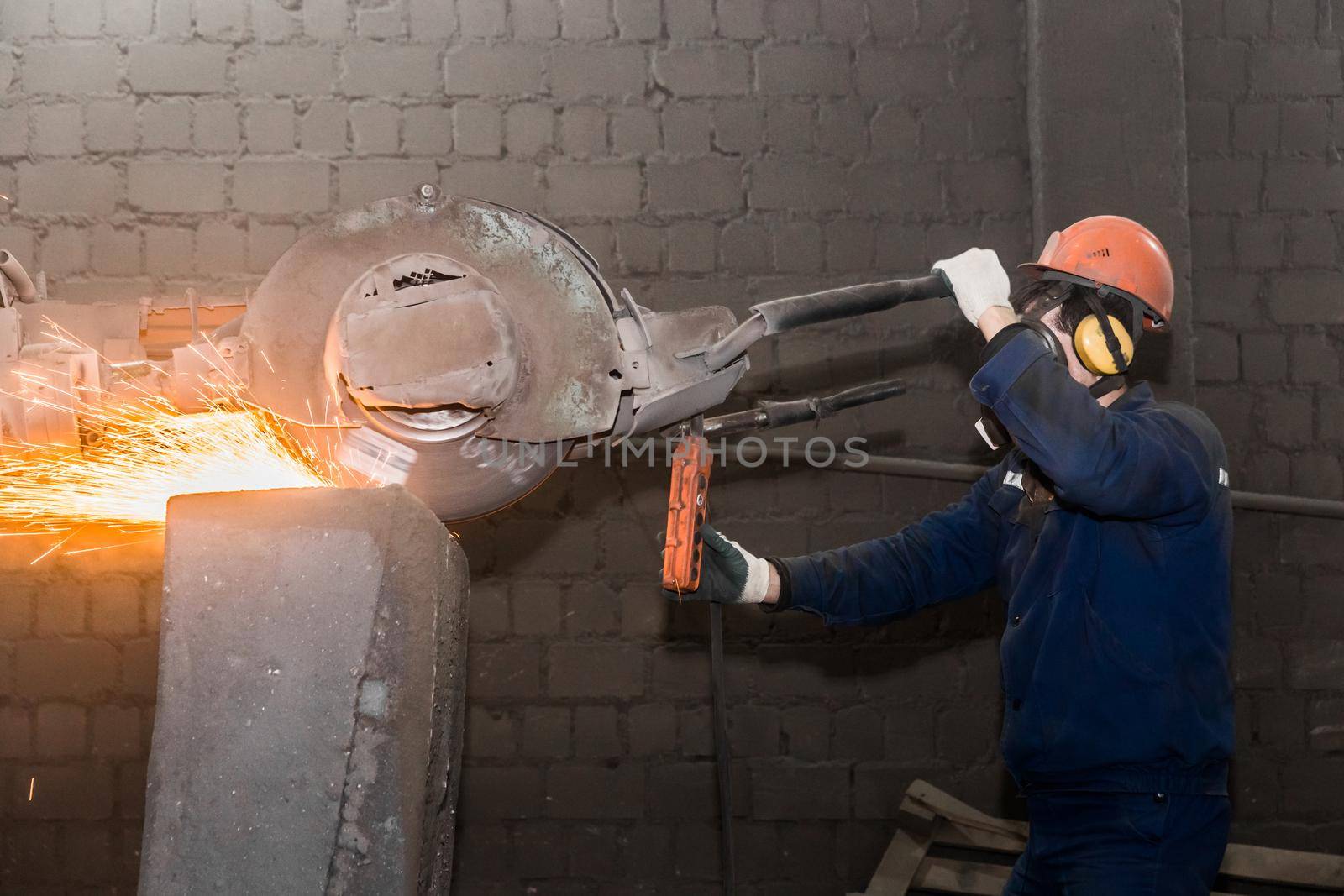 A male worker in a protective helmet, respirator, overalls manages heavy grinding equipment for cast iron concrete tubing with flying sparks in the workshop of an industrial plant.