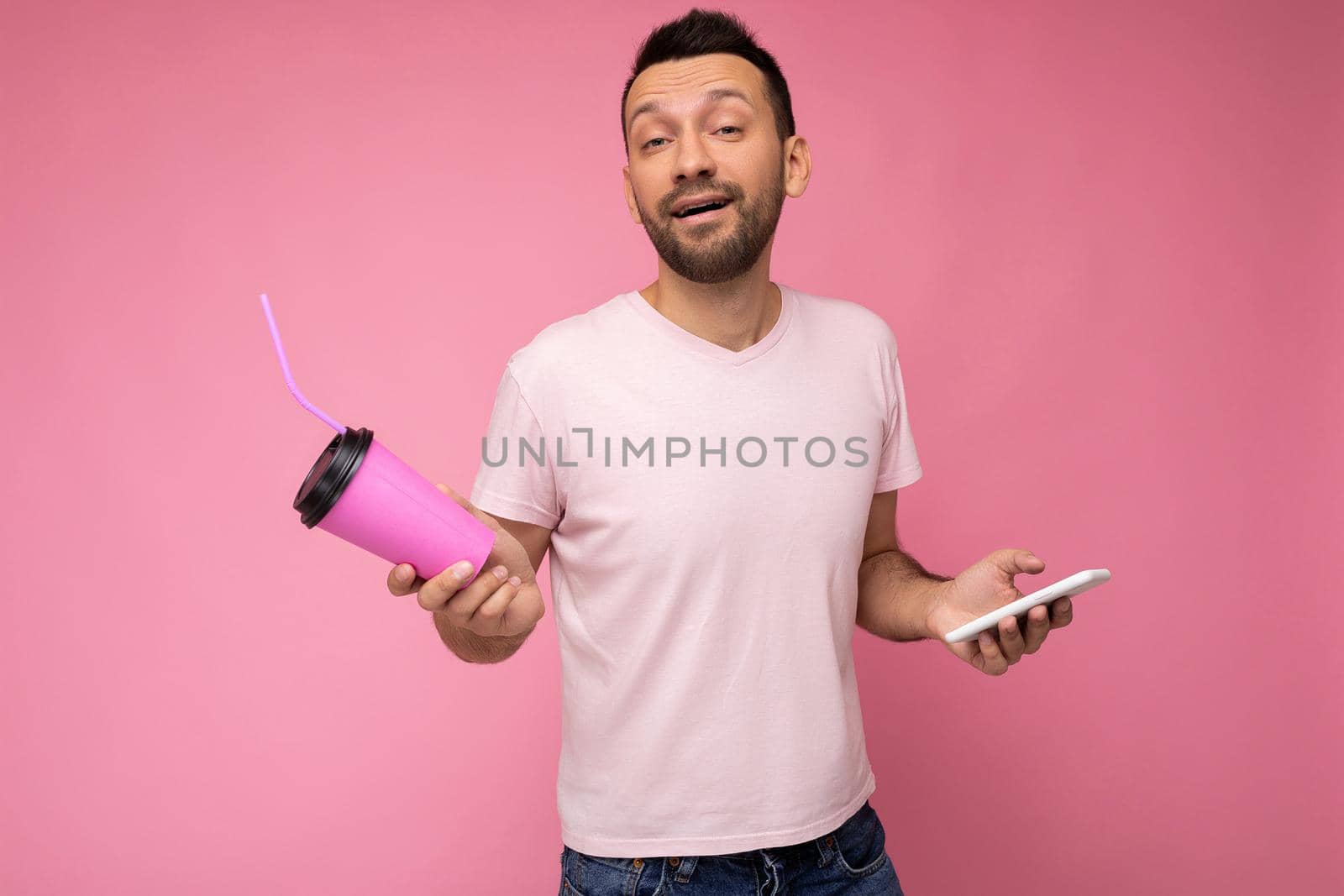 Photo of self-confident handsome young unshaven brunette man with beard wearing everyday light pink t-shirt isolated over pink background holding and using mobile phone surfing on the internet online and drinking beverage looking at camera.