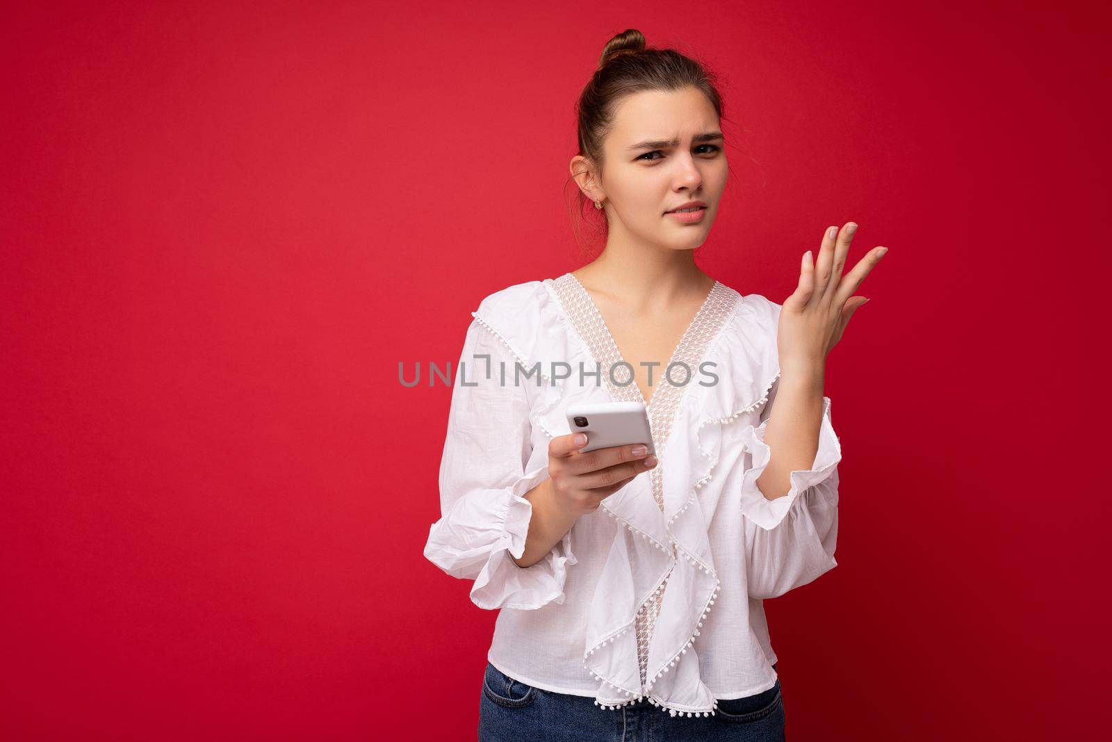 Attractive young upset brunet woman wearing white blouse standing isolated over red background reading news on the internet via phone looking at camera and having questions.