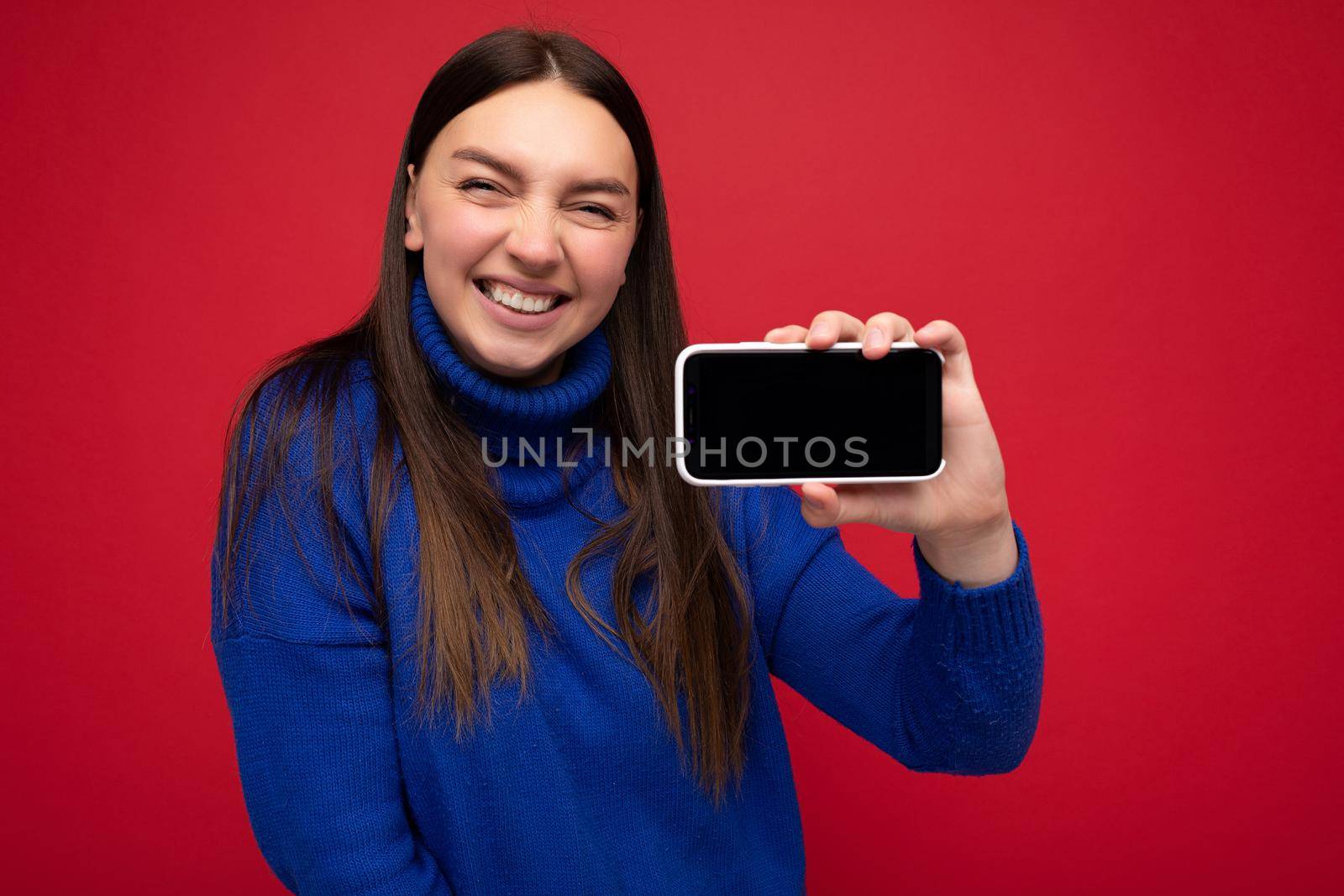 Smiling beautiful happy young brunette woman wearing casual blue sweater isolated over red background with empty space holding in hand mobile phone and showing smartphone with empty screen for mockup looking at camera.