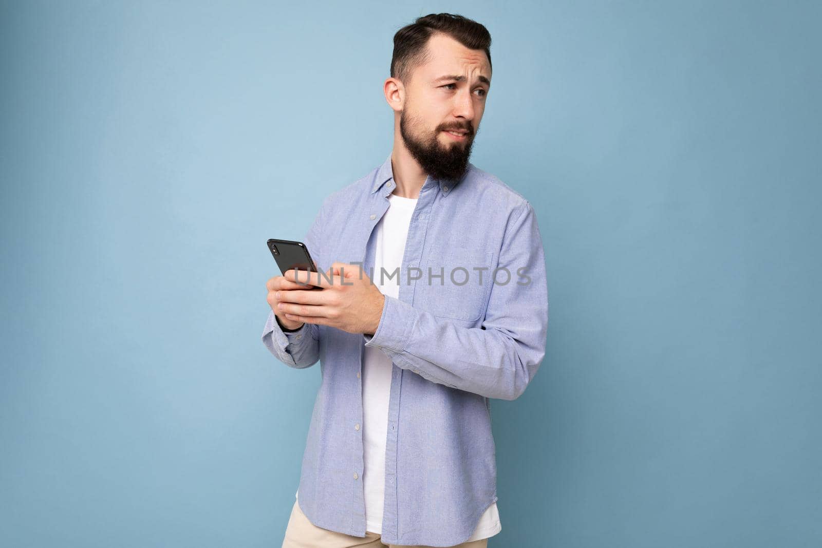 Thoughtful handsome young man isolated over background wall wearing everyday clothes holding and using mobile phone writing sms looking to the side and thinking.