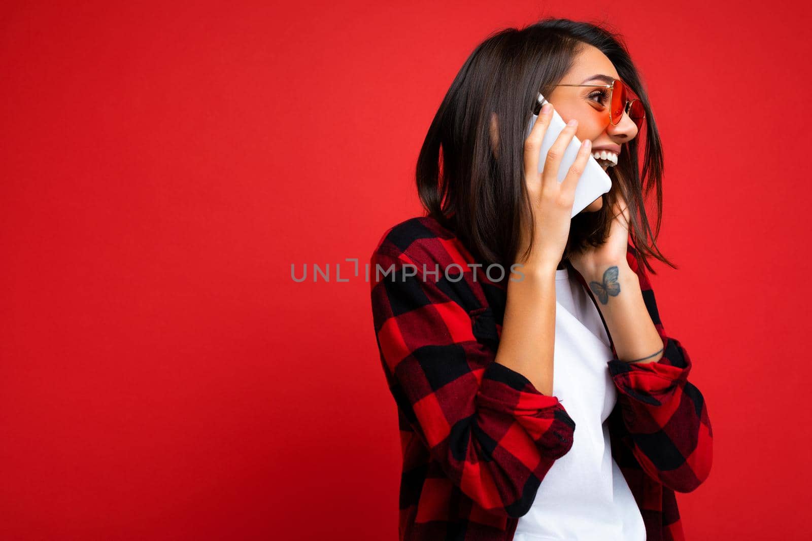 Side-profile photo of Beautiful positive young brunette woman wearing stylish red shirt white t-shirt and red sunglasses isolated over red background talking on mobile phone looking to the side.
