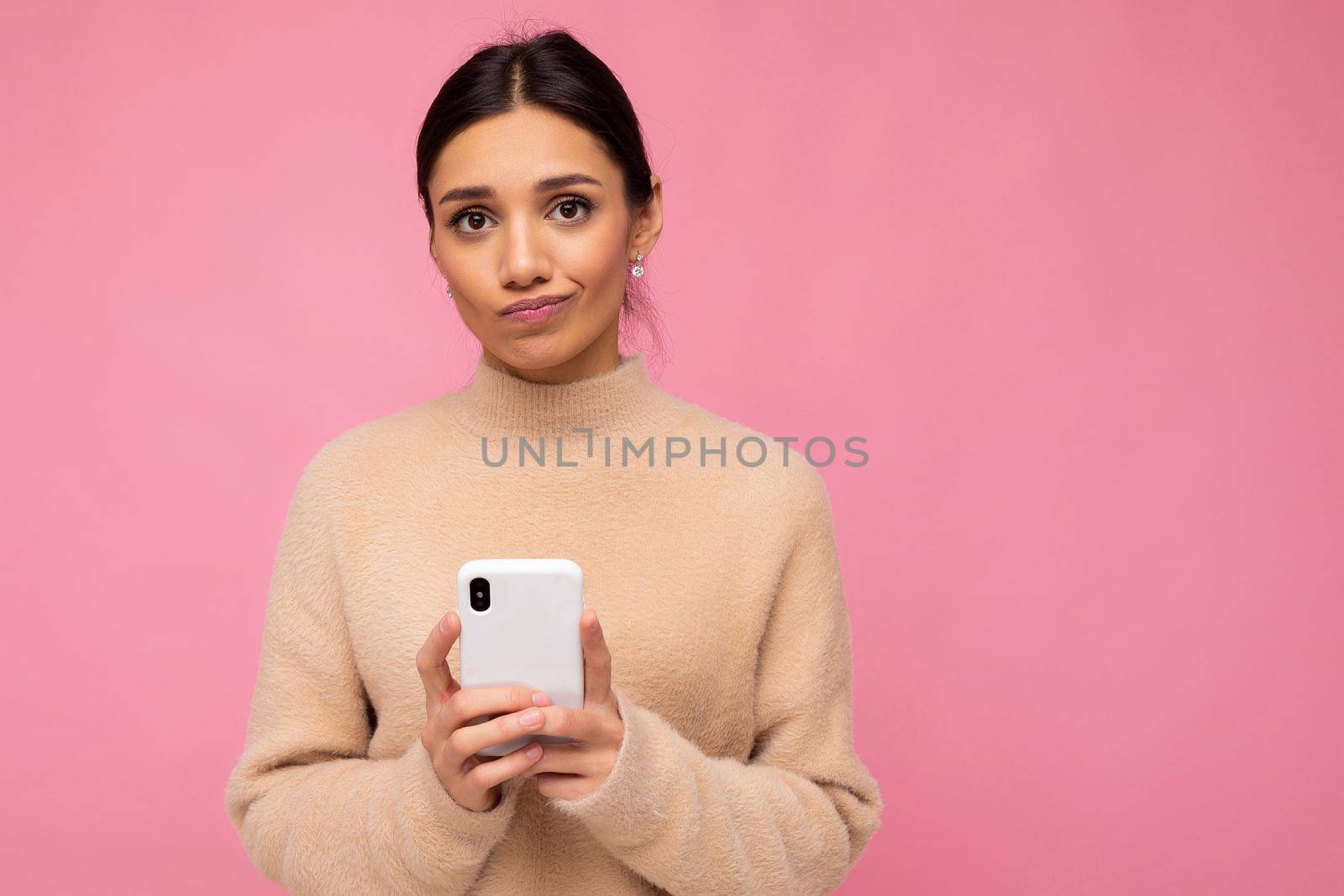 Attractive cute unhappy young brunet woman wearing beige warm sweater standing isolated over pink background surfing on the internet via phone looking at camera.