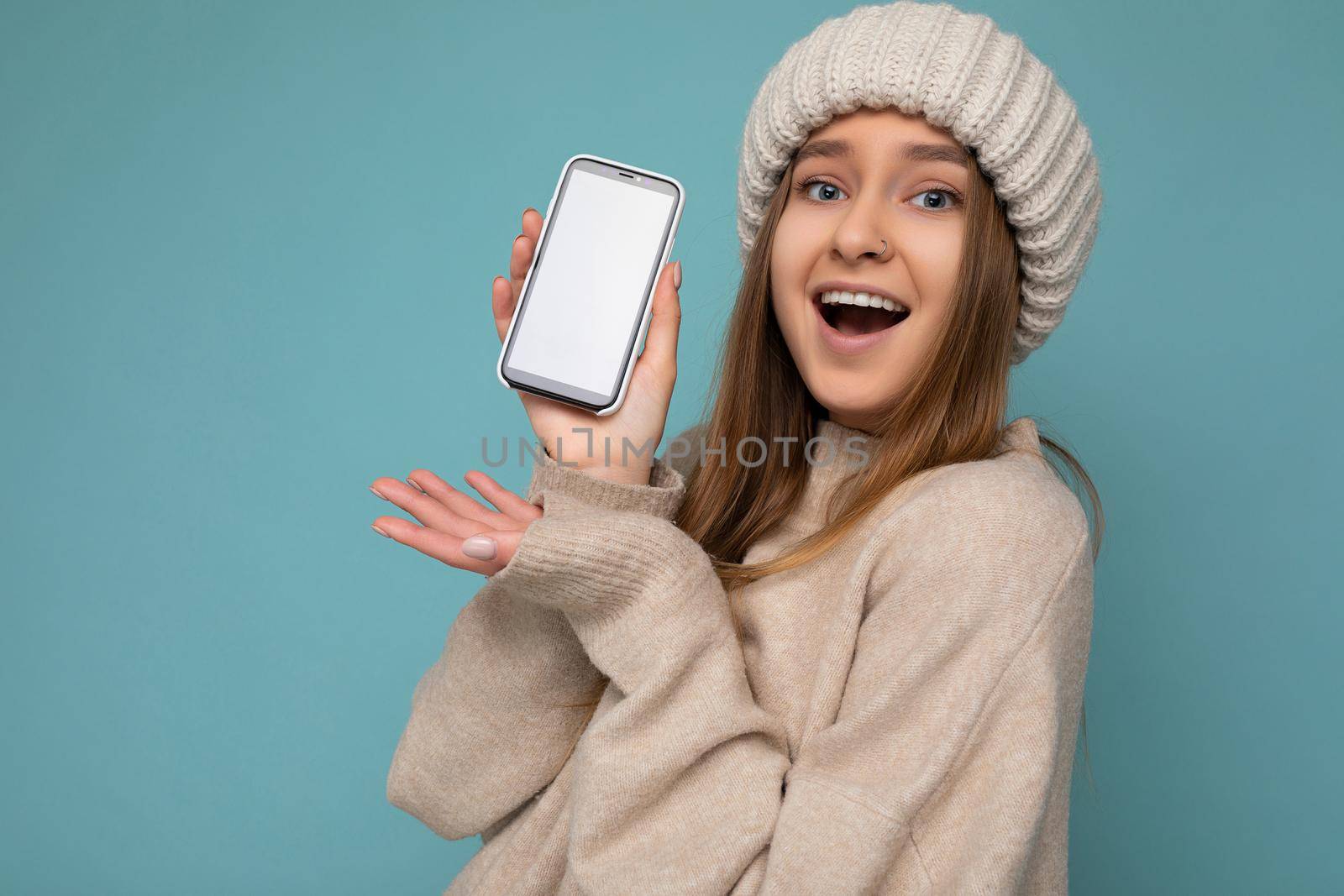 Closeup Photo of amazed beautiful positive good looking young woman wearing stylish beige sweater and beige knitted winter hat poising isolated over blue background with empty space holding in hand and showing mobile phone with empty screen for mockup looking at camera.
