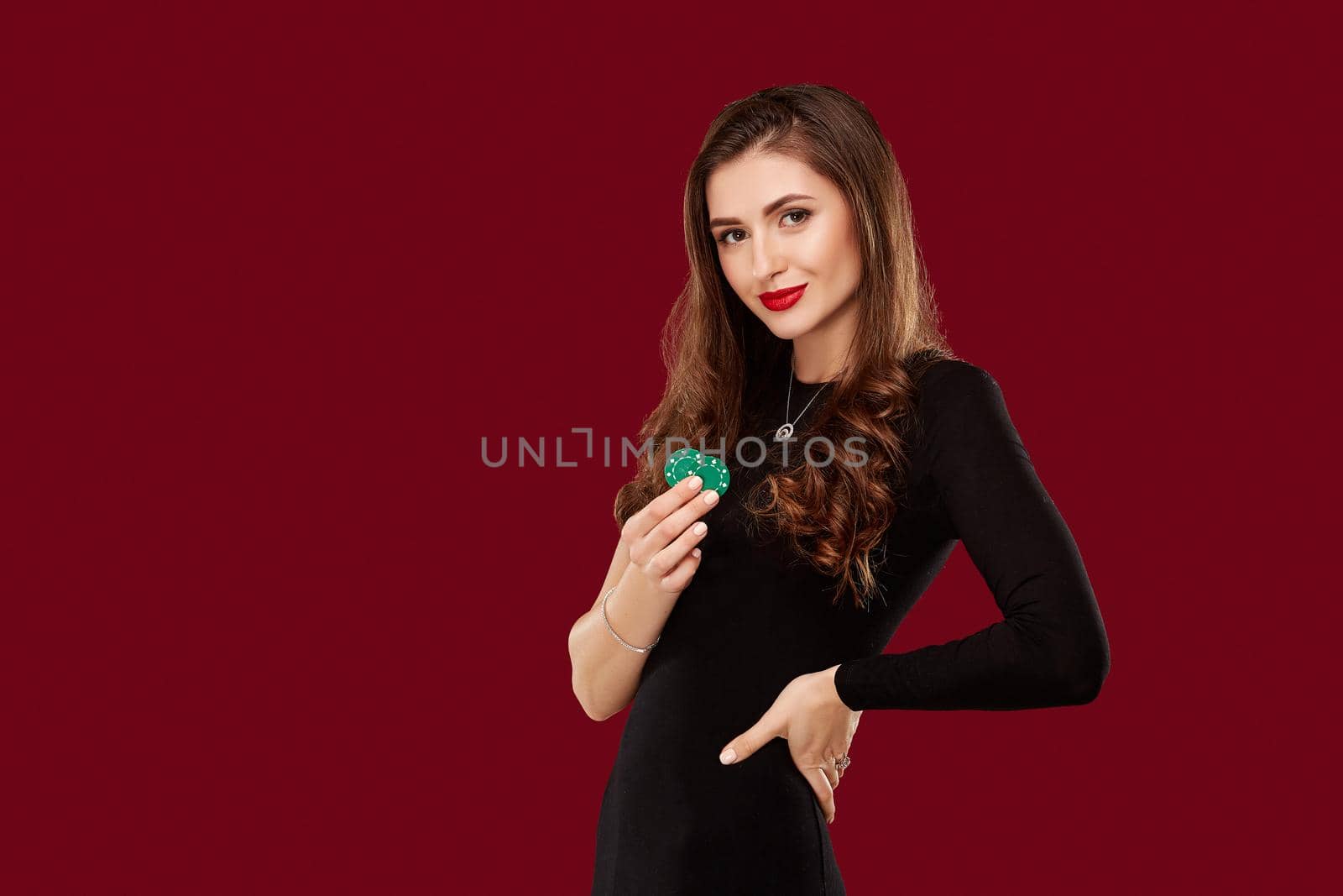 Pretty long hair woman in black dress holding chips for gambling in casino. Studio shot on red background. Poker. Two green Chips in Hand