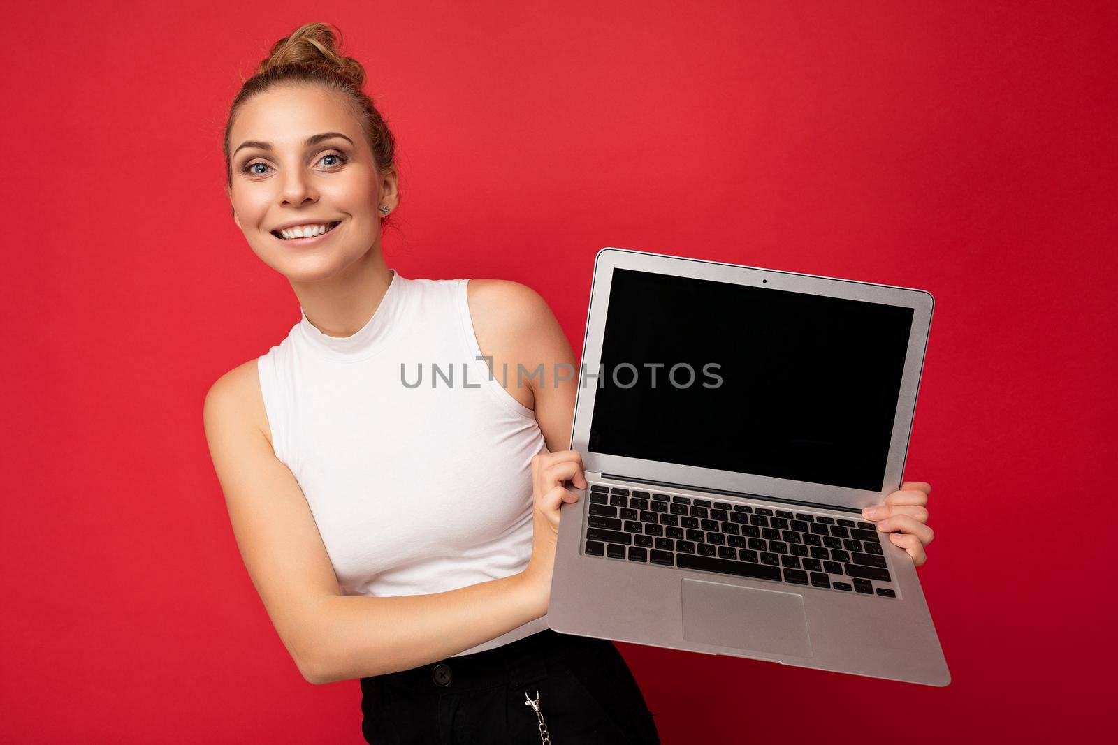 beautiful smiling fascinating happy blond young woman with gathered hair looking at camera holding computer laptop wearing white t-shirt isolated over red wall background.
