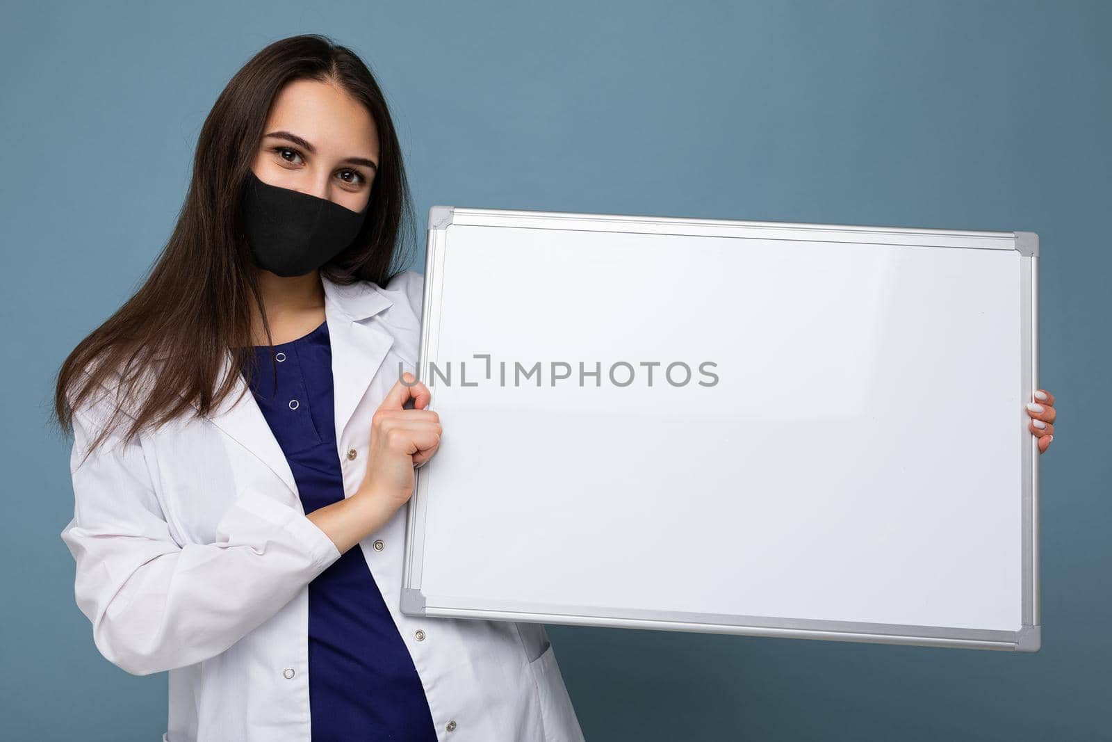 Nurse in protective face mask and white medical coat holding an empty magnetic board isolated on blue background.