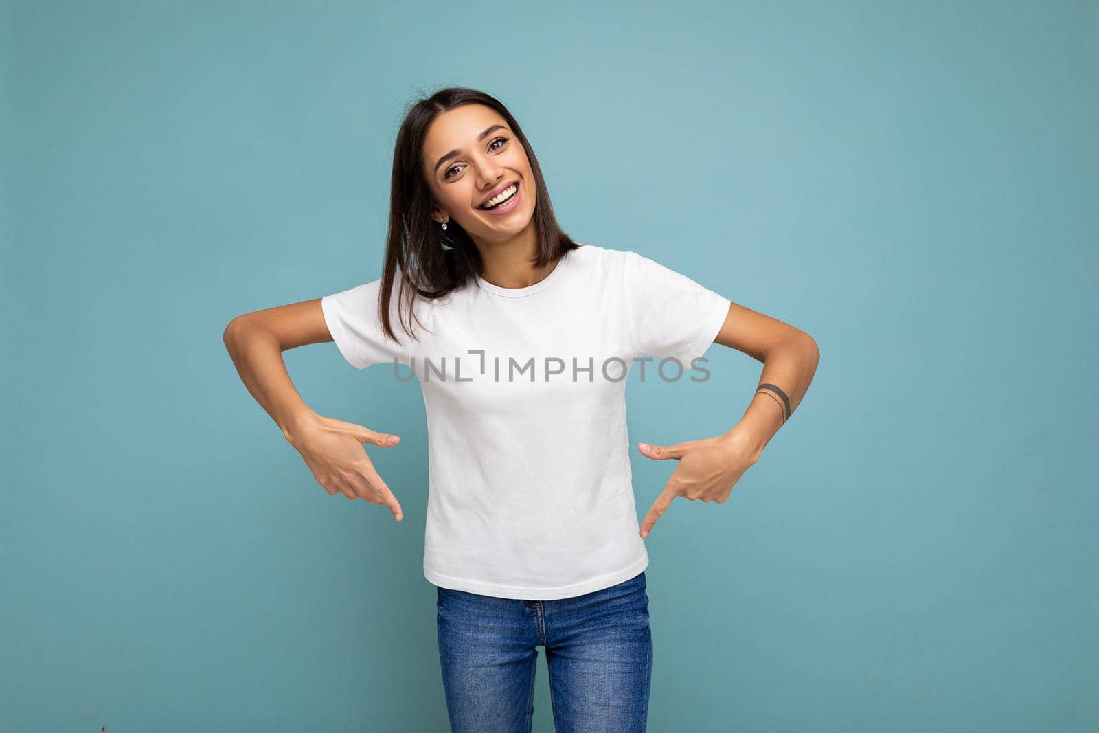 Photo of positive happy smiling young charming brunette woman with sincere emotions wearing casual white t-shirt for mockup isolated over blue background with copy space and pointing at empty space.