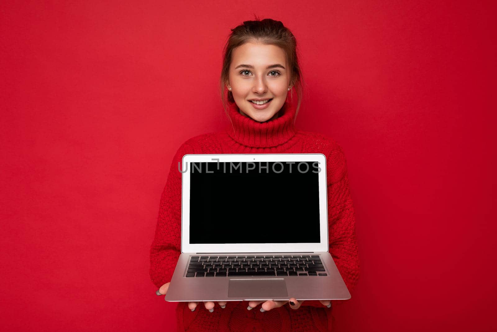Photo of Beautiful satisfied happy young woman holding computer laptop looking at camera wearing red sweater isolated over red wall background. cutout