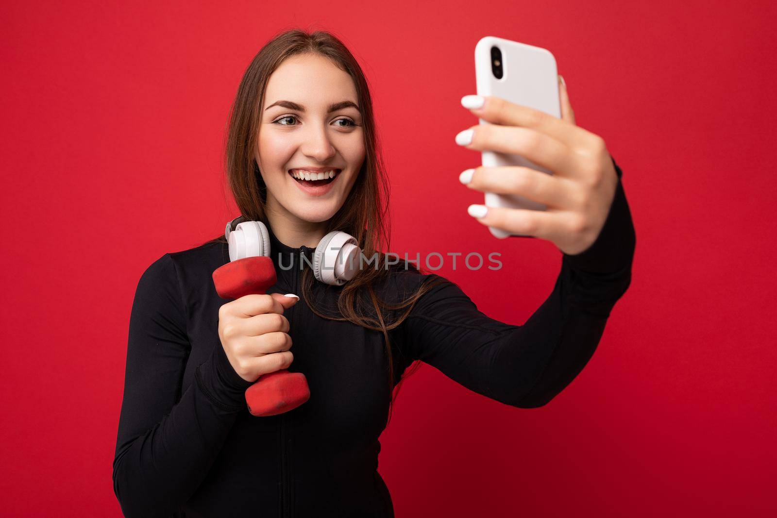 closeup Photo of beautiful smiling positive young brunette woman wearing black sport clothes white headphones nad holding red dumbbell standing isolated over red background using mobile phone taking selfie photo looking at gadjet display.