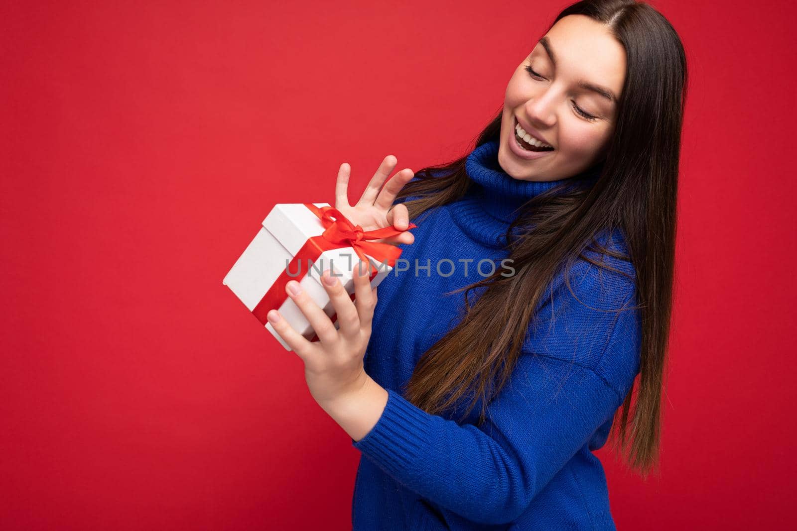 Shot of attractive happy smiling young brunette woman isolated over red background wall wearing blue casual sweater holding white gift box with red ribbon and looking at camera.
