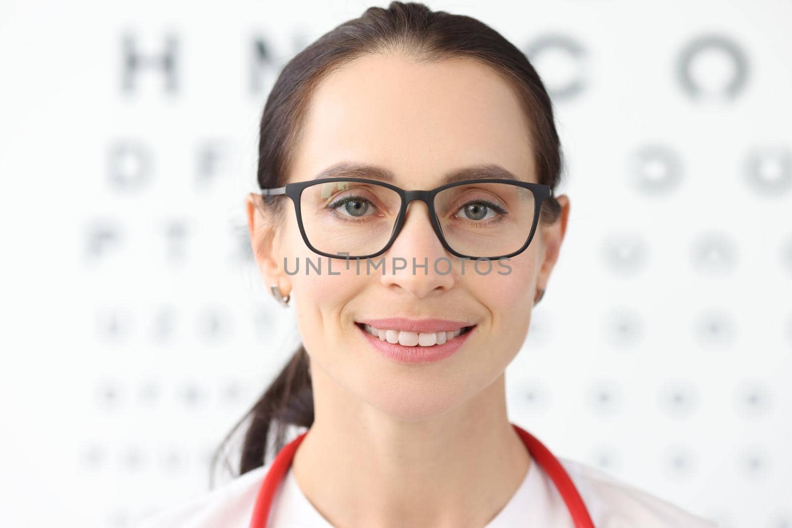 Woman ophthalmologist with glasses standing against background of table for eye test by kuprevich