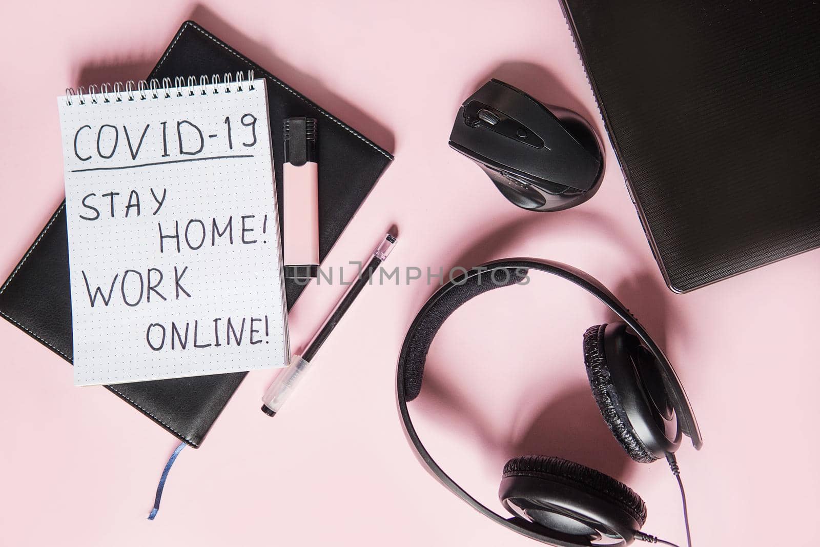 Top view of a desktop with notebooks, headphones, pens, and a laptop.Text Covid-19, stay at home, WORK ONLINE. Work from home using an online device, protection from coronavirus during the quarantine period. Pink background.
