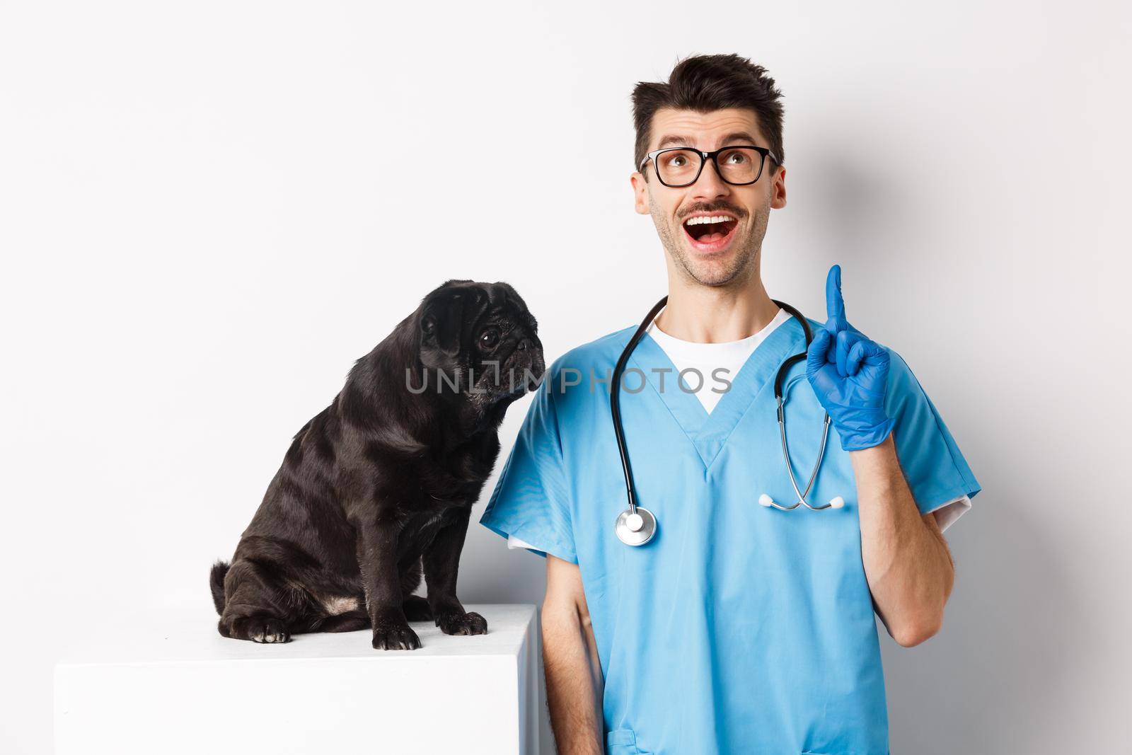 Handsome young doctor at vet clinic pointing finger up and looking amazed, standing near cute black pug dog, white background by Benzoix