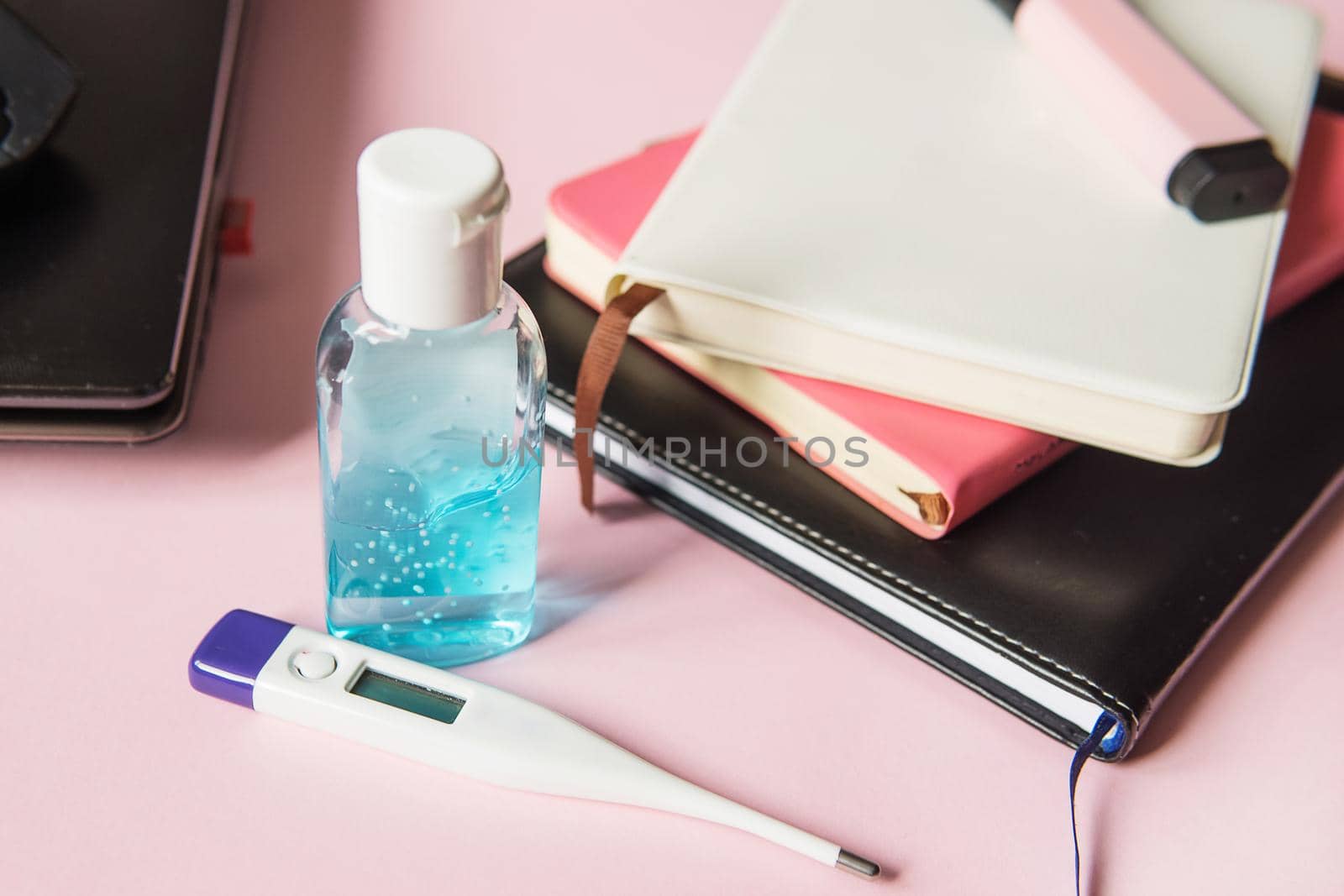 On the desktop, a close-up of a thermometer and hand gel, as well as notebooks, a marker and a laptop. Work from home using an online device protection from coronavirus during the quarantine period. Pink background. Selective focus.