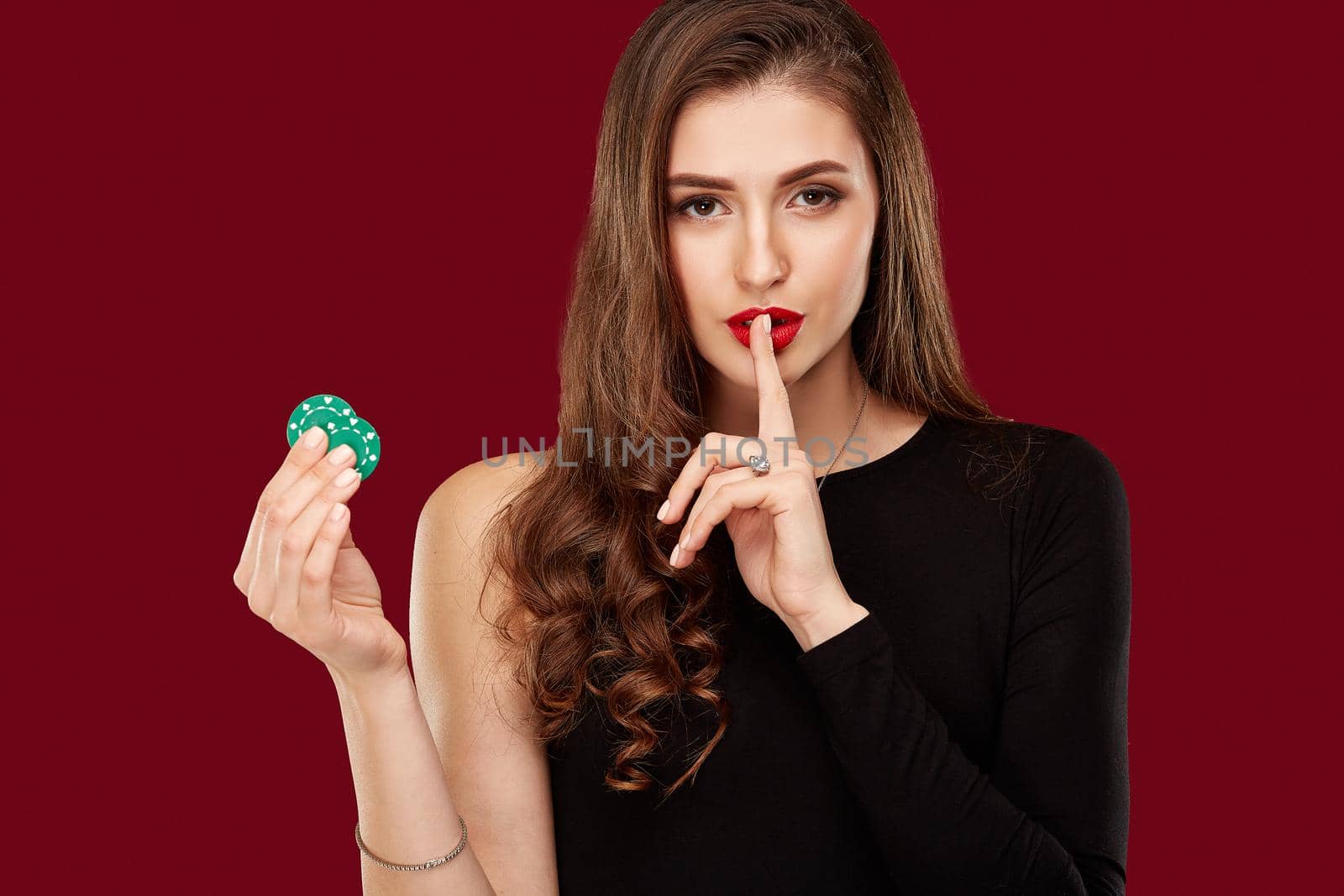 Pretty long hair woman in black dress holding chips for gambling in casino. Studio shot on red background. Poker. Two green Chips in Hand