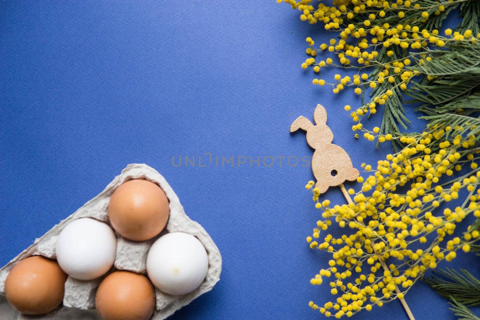 Easter background, eggs and a wooden hare on a blue background, decorated with Mimosa flowers, flatlay, top view, empty space for text. happy Easter.