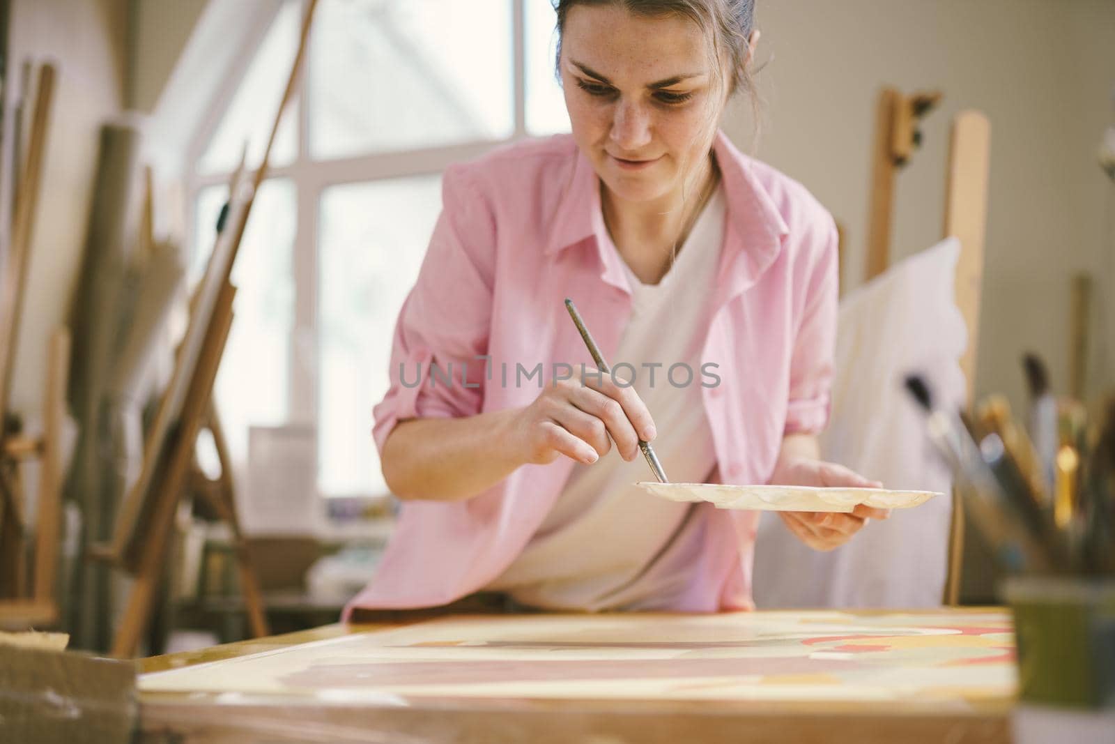 Cute woman paints on canvas in an art workshop. Artist creating picture. Art school or studio. Work with paints, brushes and easel. Hobby and leisure concept. Woman painter at workspace by Tomashevska