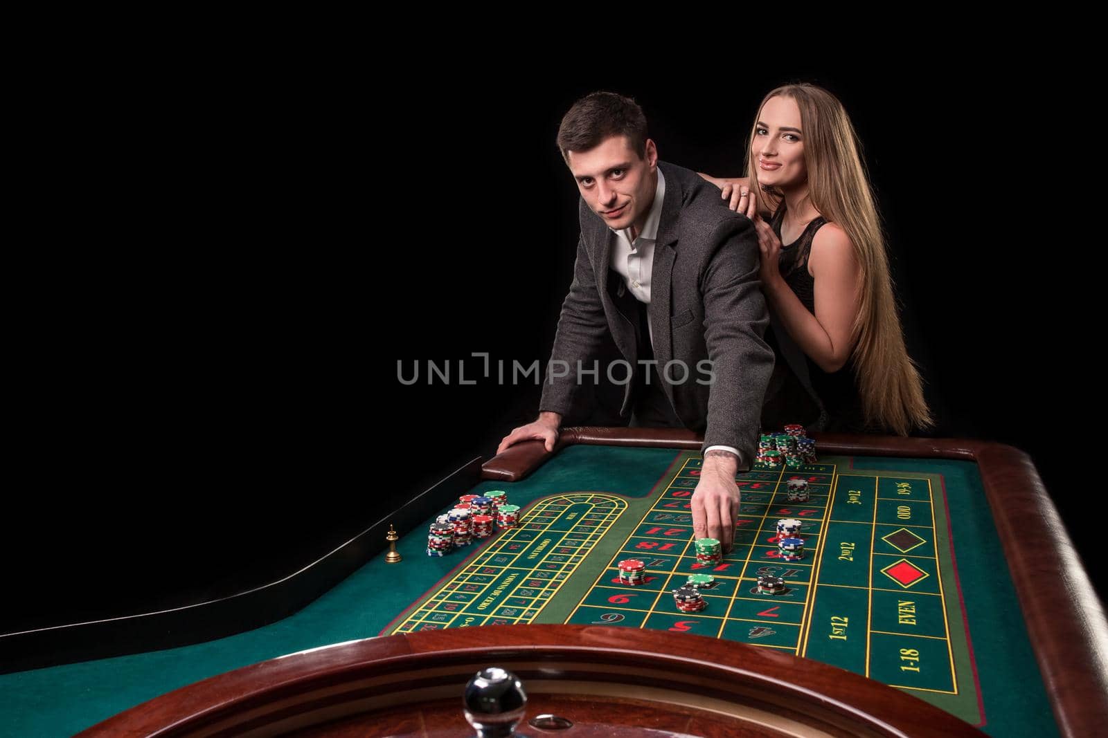 Elegant couple at the casino betting on the roulette, on a black background. A man in a suit with a beautiful young woman in a black dress
