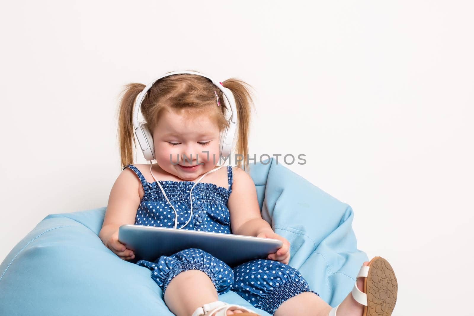 Cute little girl in headphones listening to music using a tablet and smiling while sitting on blue big bag. On white background.