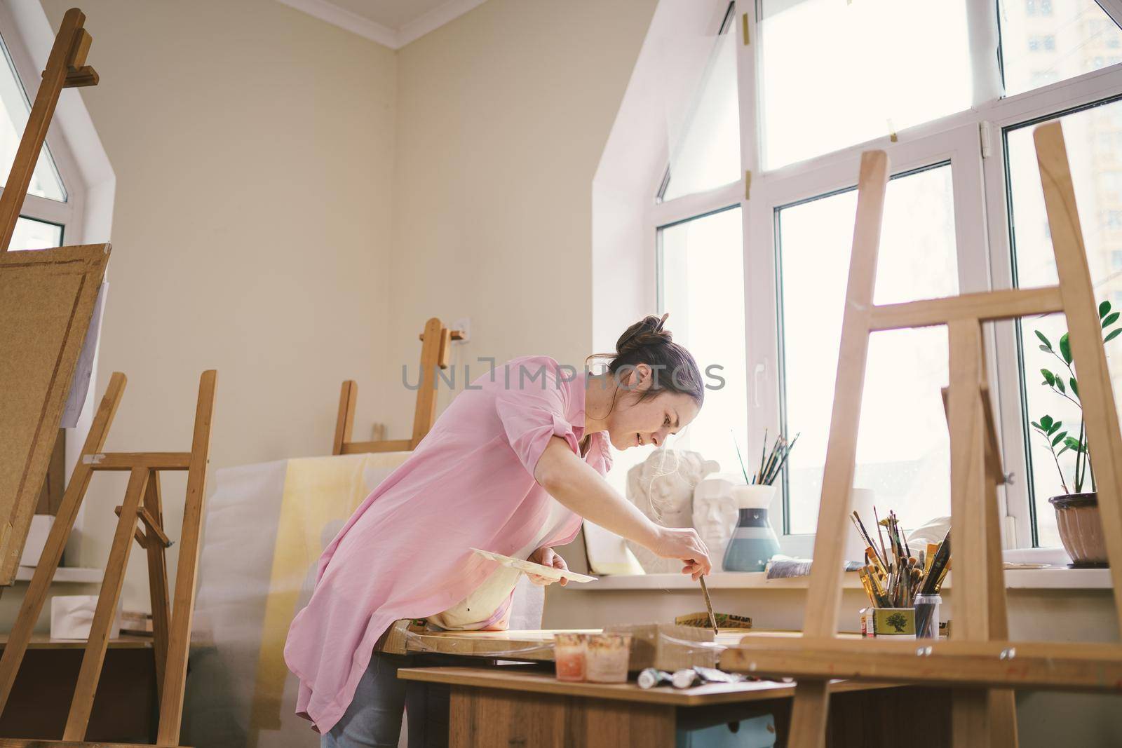 Caucasian woman artist working on a painting in bright daylight studio. Happy artist draws an art project with paints and a brush in the workshop. Hobby. Artist at work. Creative profession by Tomashevska
