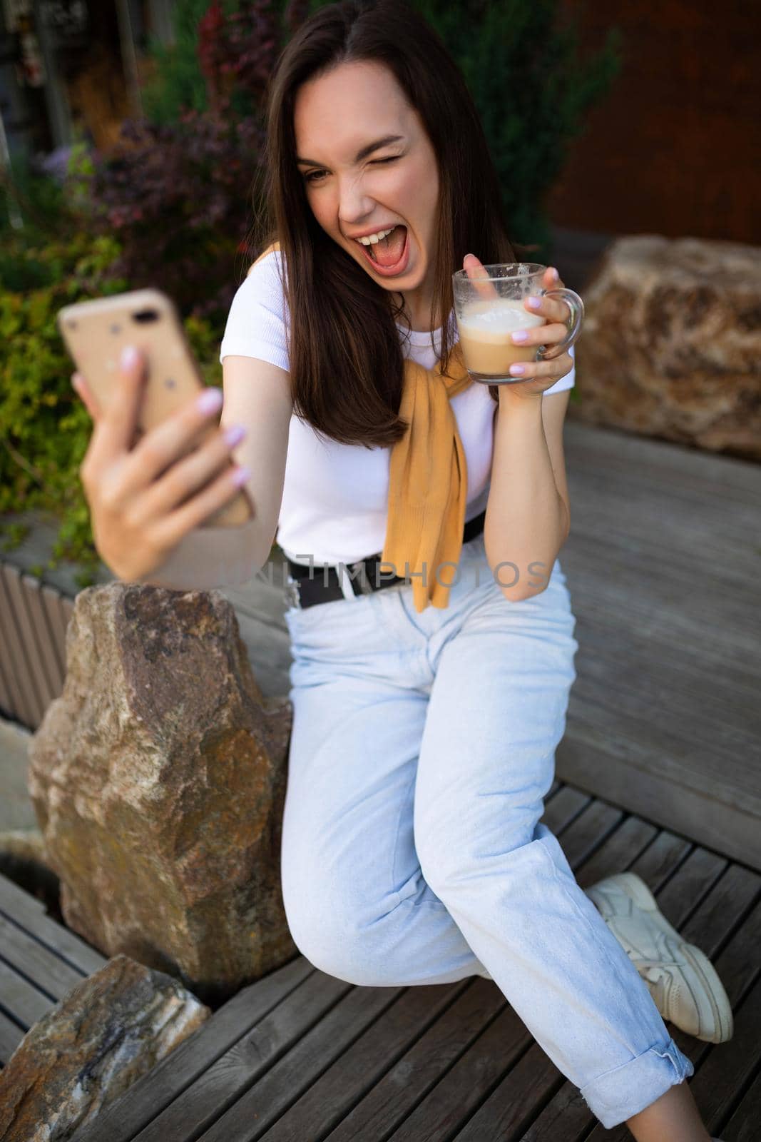 free young woman drinking coffee and chatting in a smartphone while sitting on a bench in a summer park.