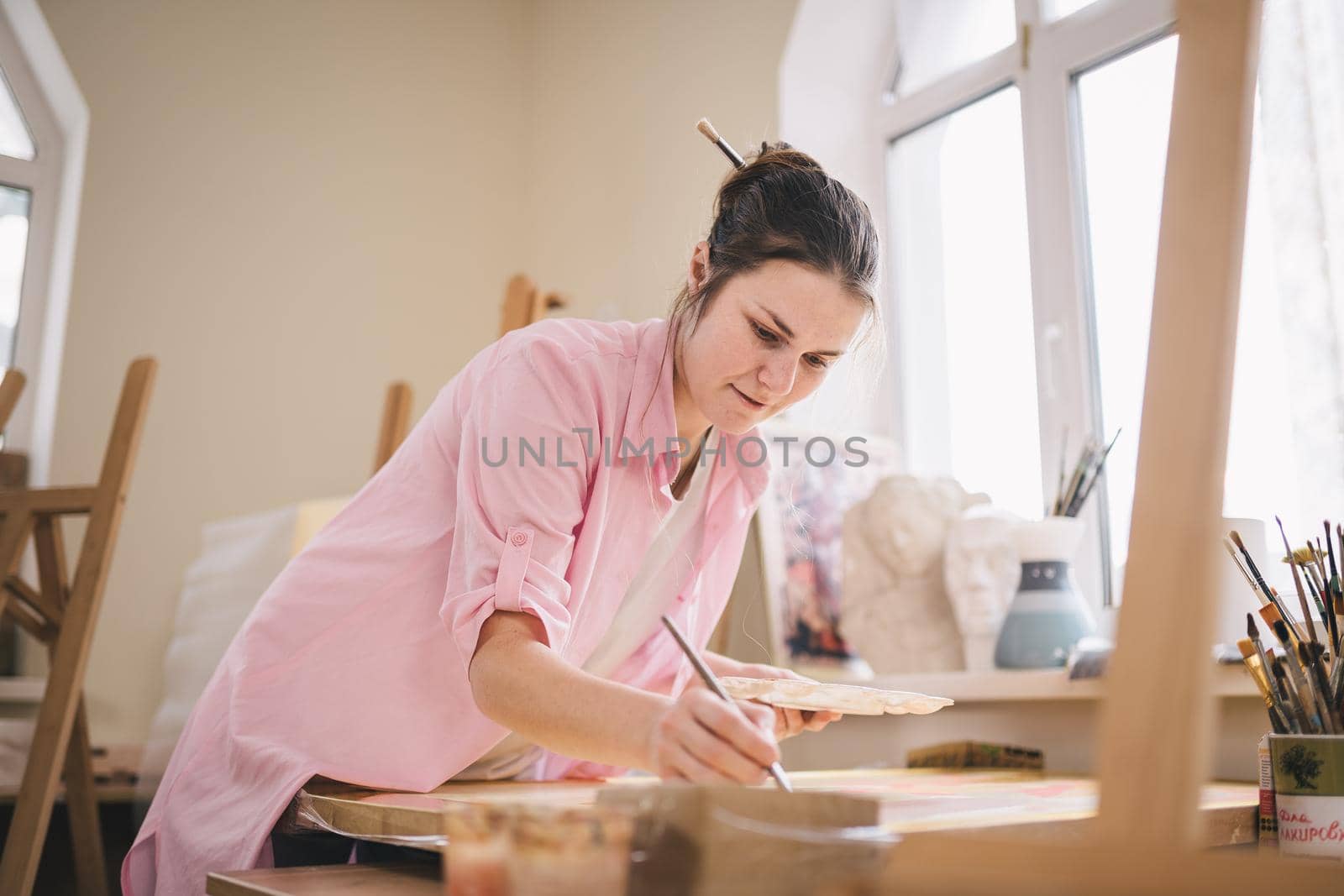 Caucasian woman artist working on a painting in bright daylight studio. Happy artist draws an art project with paints and a brush in the workshop. Hobby. Artist at work. Creative profession by Tomashevska