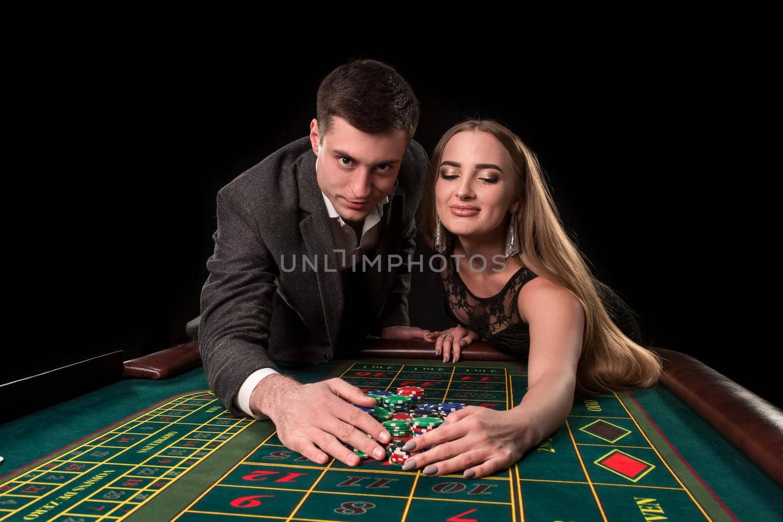 Young beautiful couple takes their winnings at the roulette table at the casino, on a black background. A man in a suit with a woman in a black dress