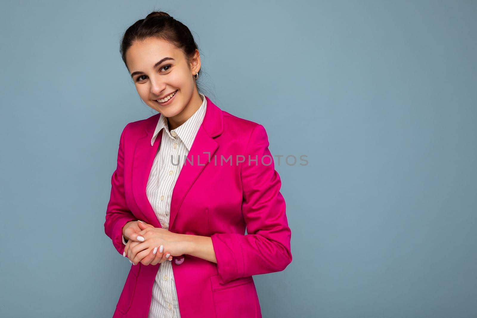 Portrait of young attractive beautiful positive smiling brunette woman with sincere emotions wearing casual white shirt and trendy pink jacket isolated over blue background with free space.