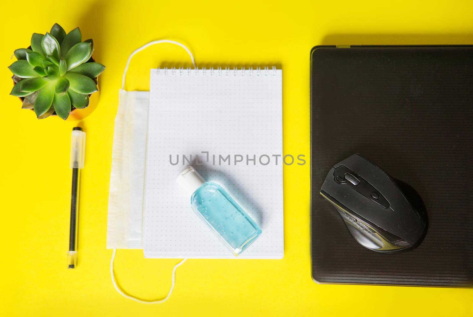 Top view of a desktop with notebooks, pens, and a laptop. Work from home using an online device, protection from coronavirus during the quarantine period. Protective masks and antiseptic on a yellow background.