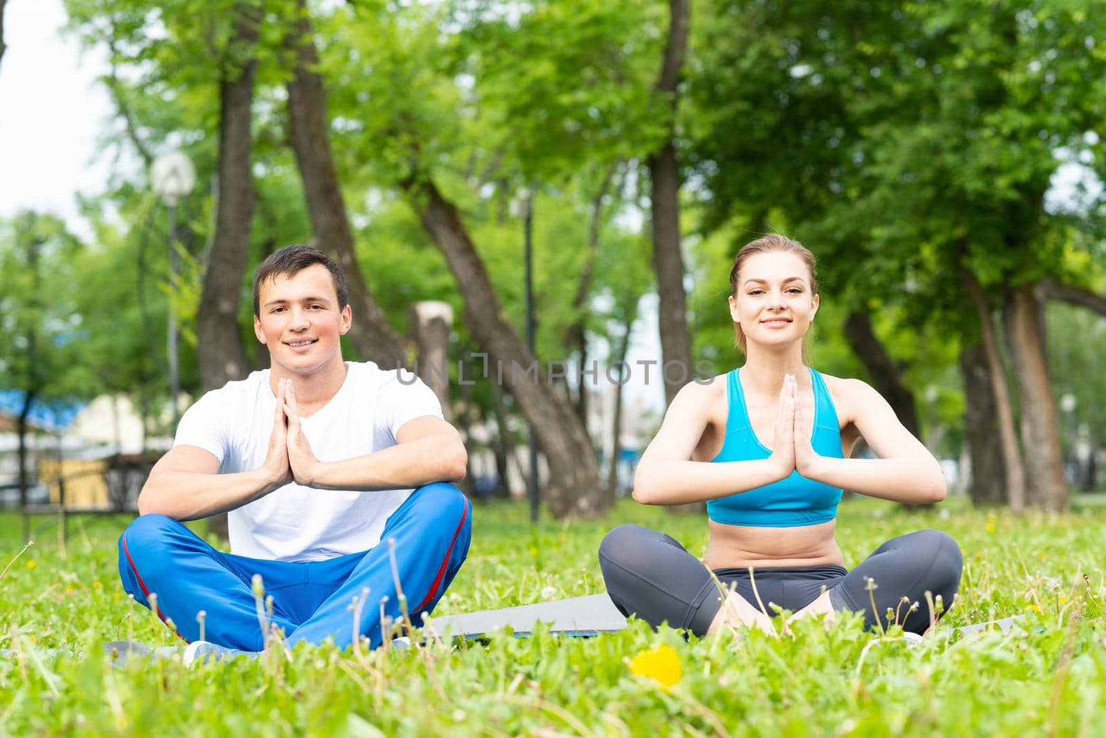 Young couple doing yoga in park together. Man and woman sitting in yoga pose on green grass. Training and meditation in yoga class outdoor at sunny summer day. Morning exercises and healthy lifestyle.