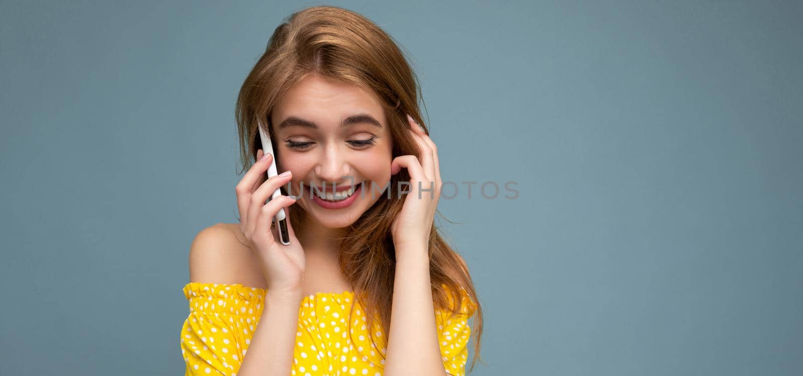 Attractive positive smiling young blonde woman wearing stylish yellow summer dress standing isolated over blue background holding and talking on mobile phone looking to down by TRMK