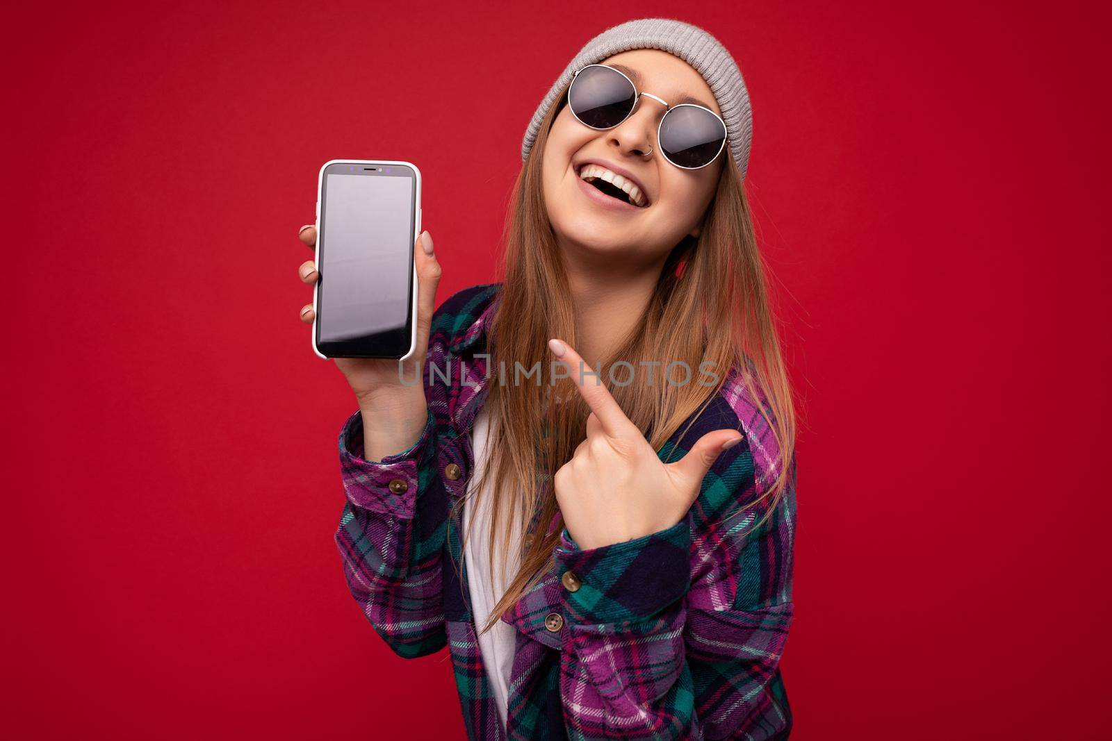 Photo of beautiful smiling young woman good looking wearing casual stylish outfit standing isolated on background with copy space holding smartphone showing phone in hand with empty screen display for mockup pointing at gadjet looking at camera by TRMK
