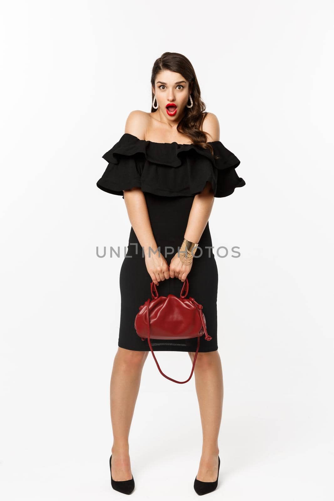 Beauty and fashion concept. Full length if silly young woman pouting and looking surprised, holding purse, wearing heels and black dress, white background.