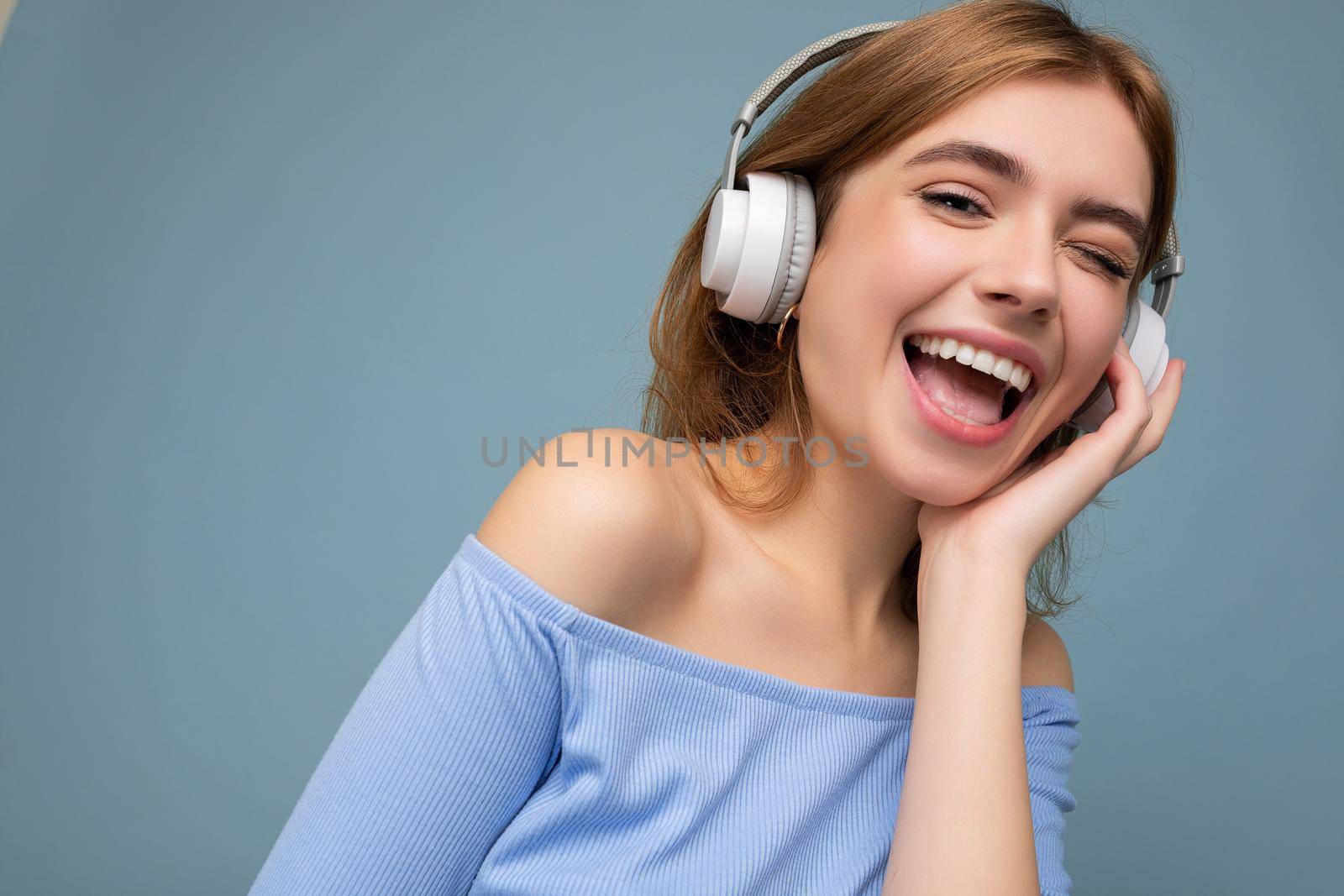 Closeup photo of attractive happy smiling young blonde woman wearing blue crop top isolated over blue background wall wearing headphones listening to music and having fun.