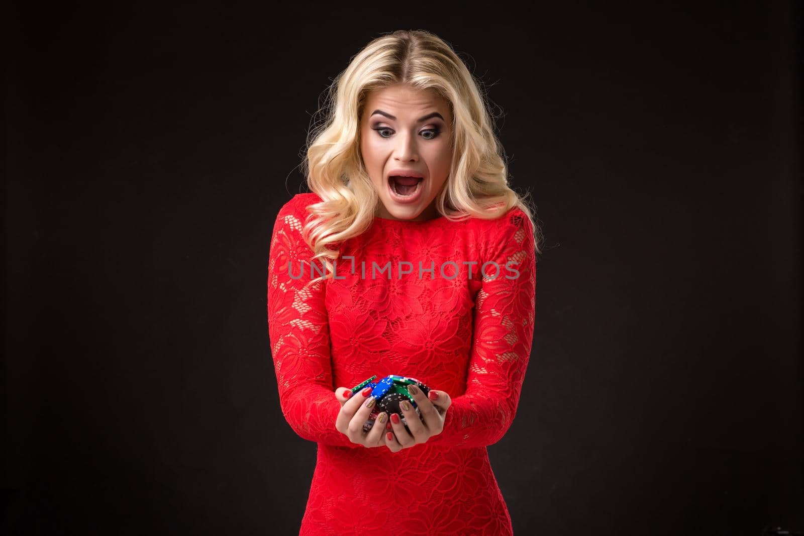 Young beautiful emotional woman with chips in hands on a black background in the studio. Portrait of a beautiful blonde in a red dress. Poker